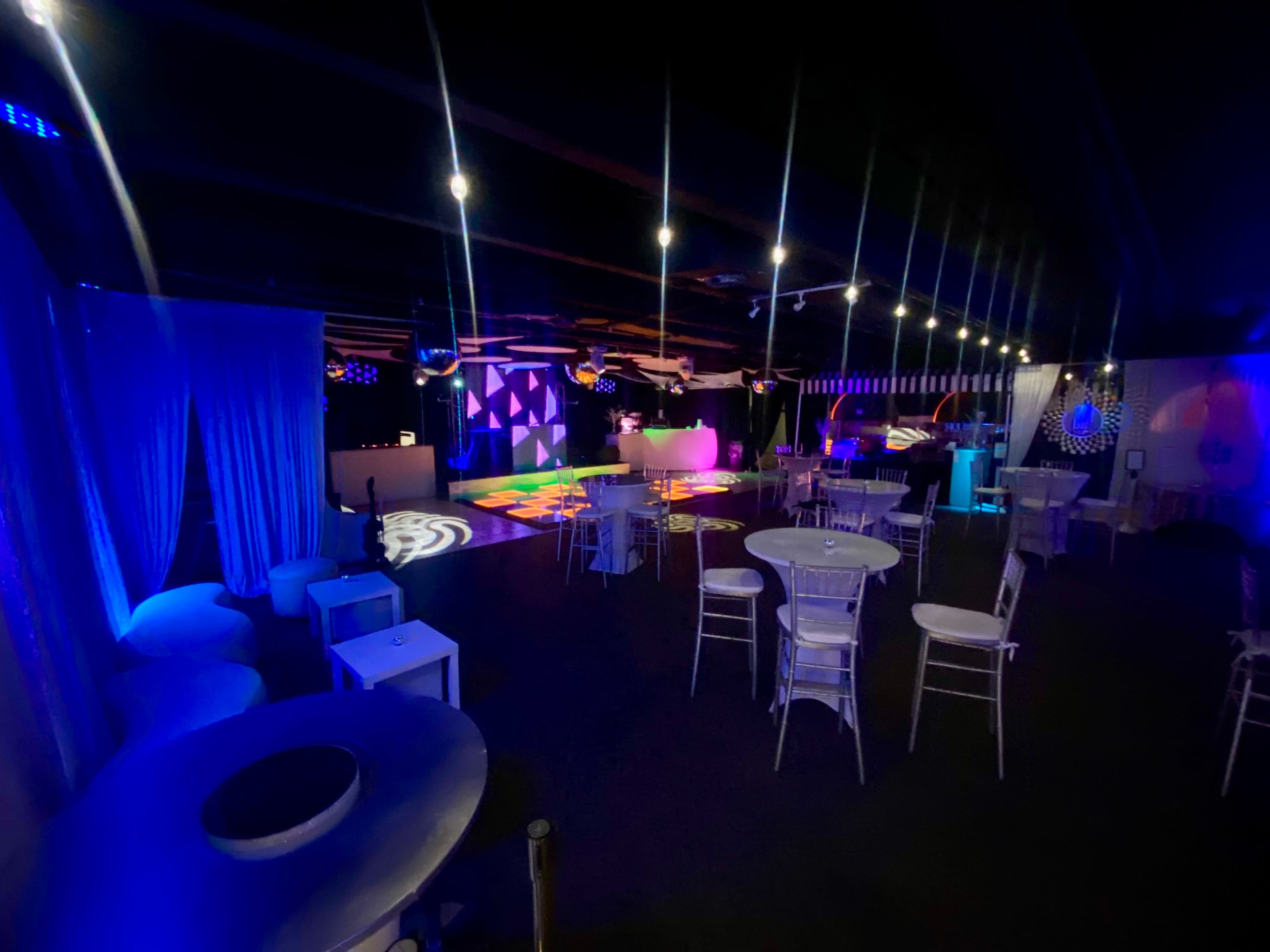 The image shows a dimly lit event space with colorful lighting, featuring white tables and chairs arranged around a dance floor.