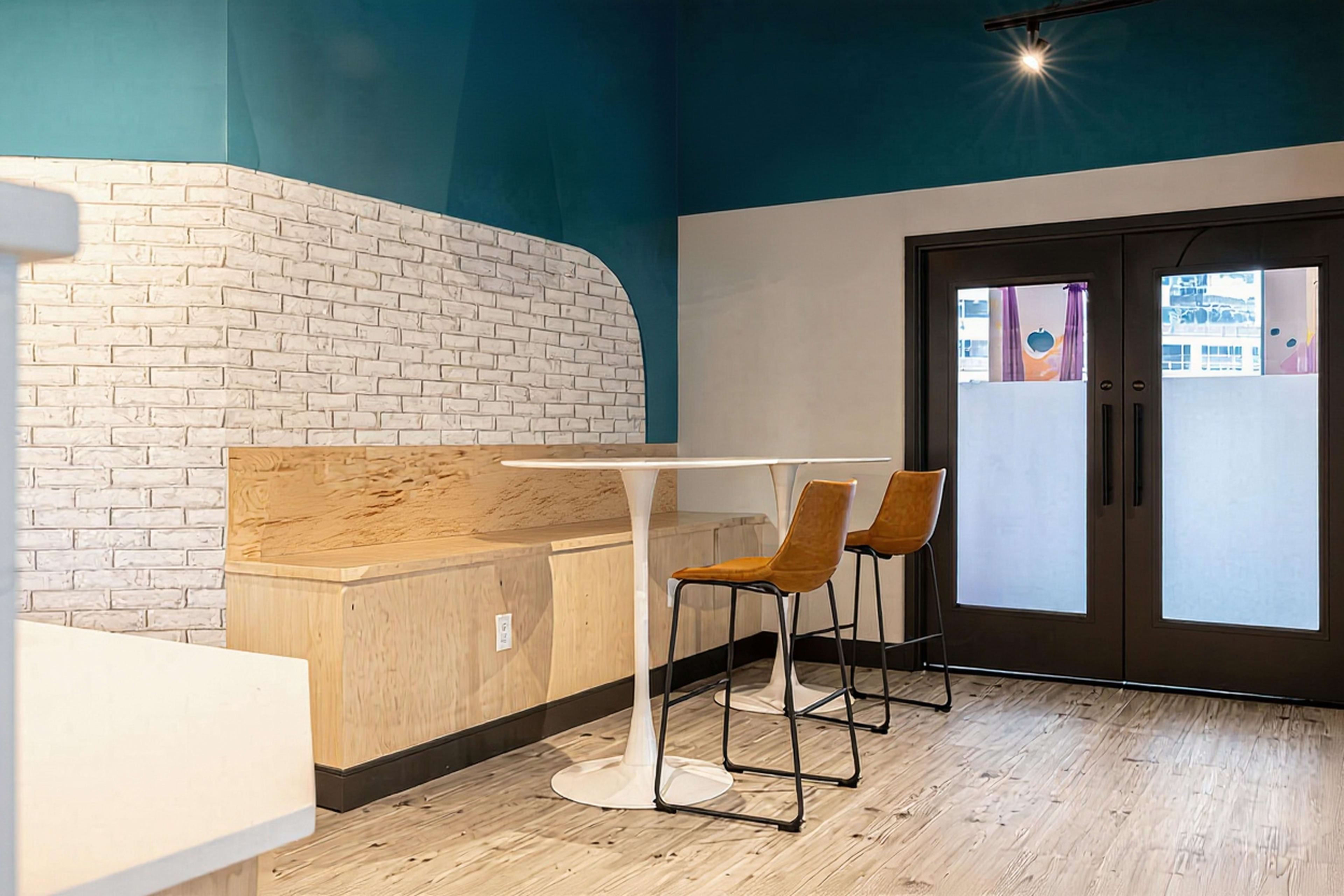 The image shows a small dining area with a white table and two brown chairs against a colorful wall and large glass doors.