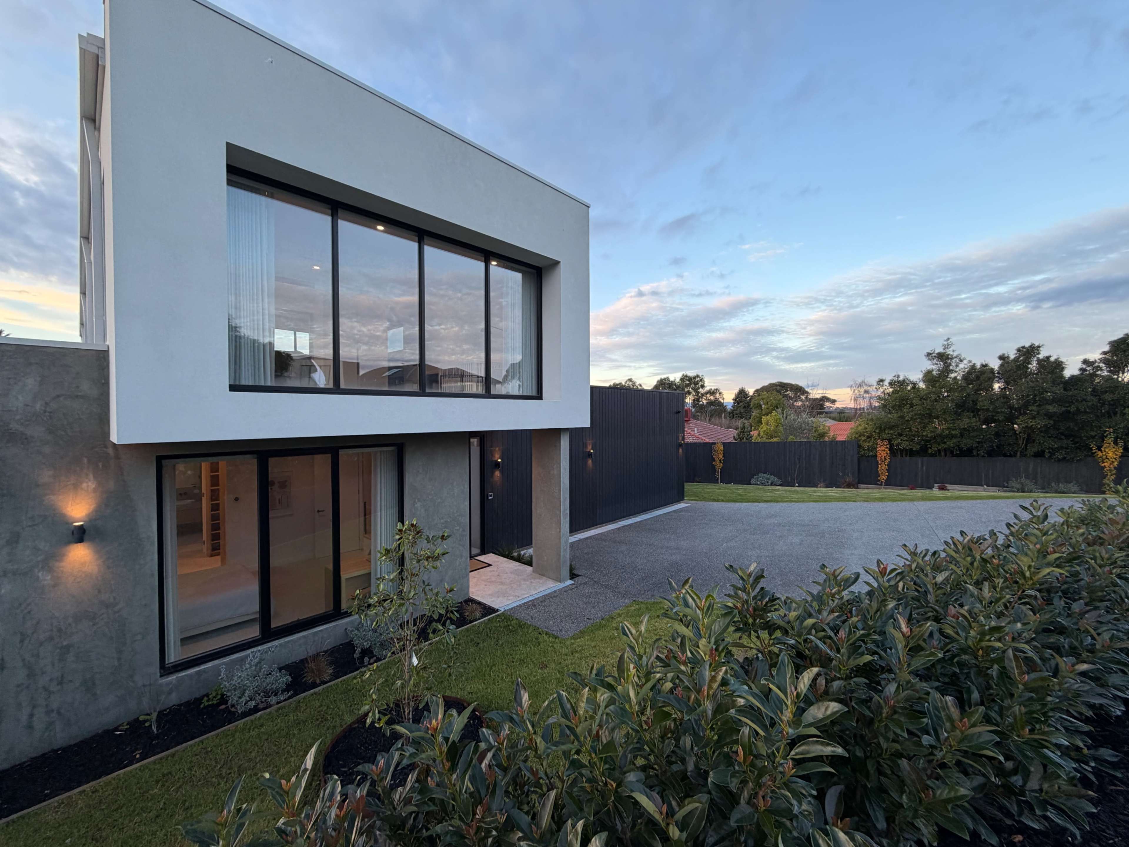 The image shows a modern multi-level house with large windows, surrounded by neatly landscaped greenery and a gravel driveway.