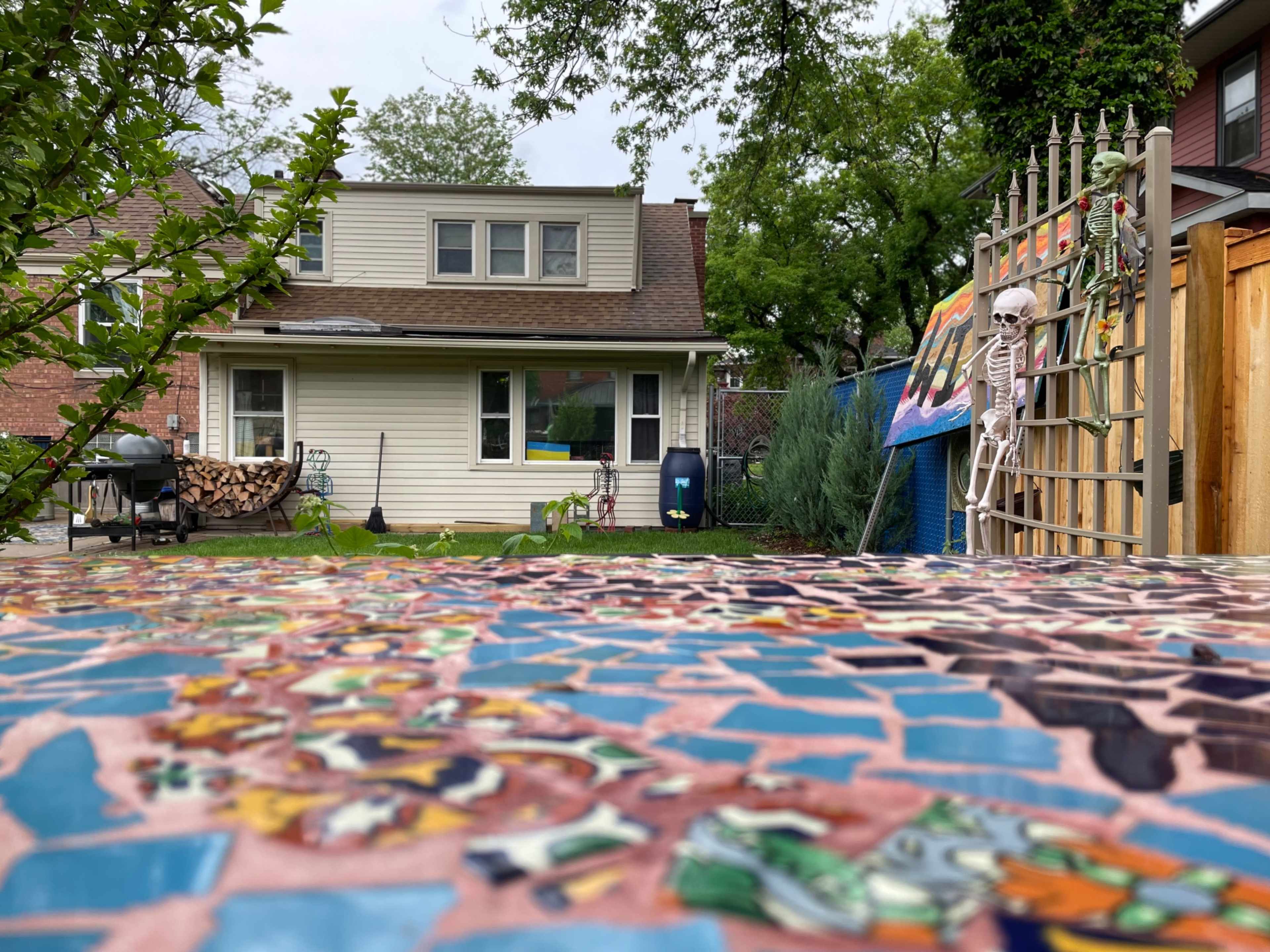A backyard with a mosaic table in the foreground, a house in the background, and a skeleton decoration near a gate.