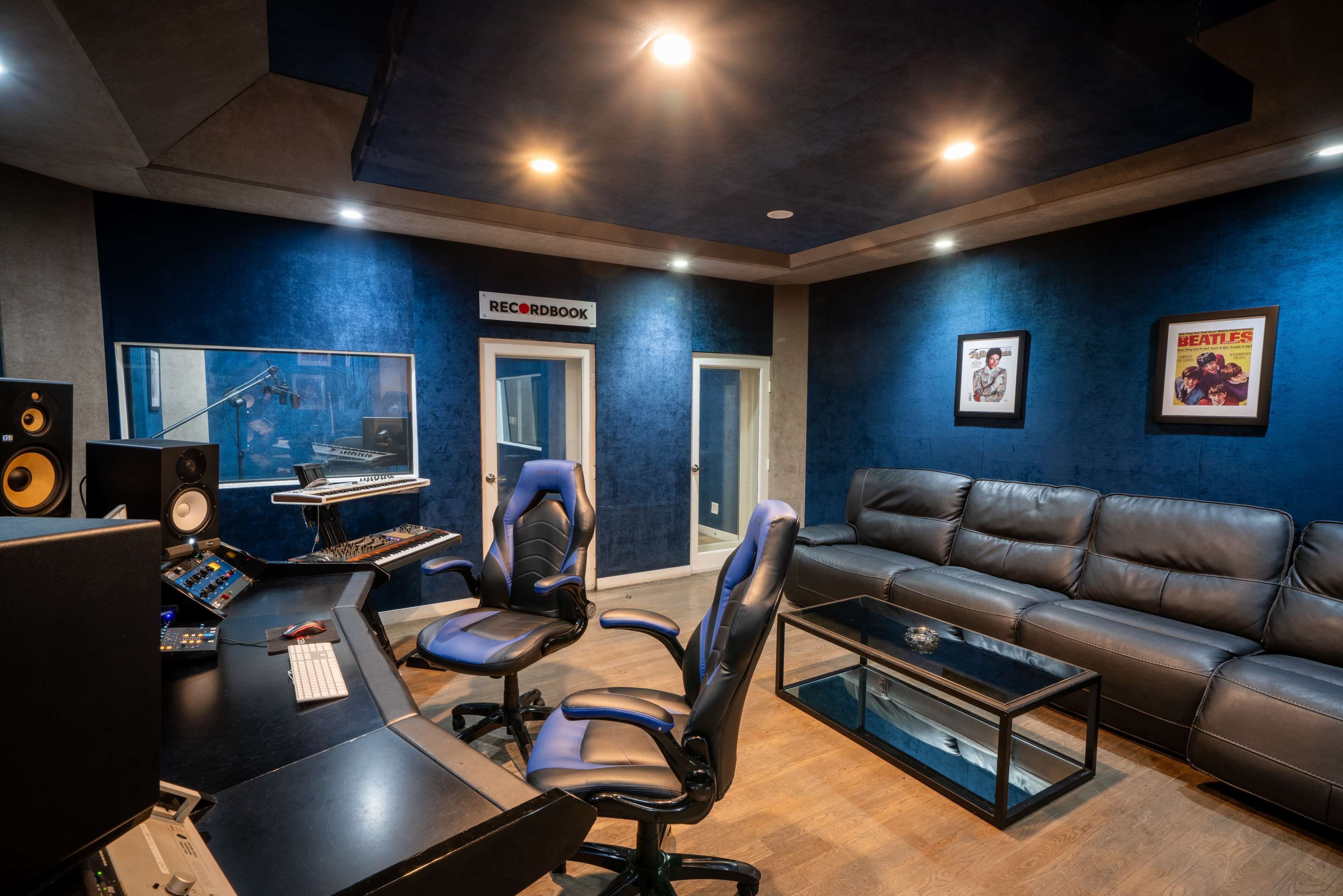 The image shows a modern recording studio with a control desk, two black ergonomic chairs, a leather couch, and blue acoustic panels on the walls.
