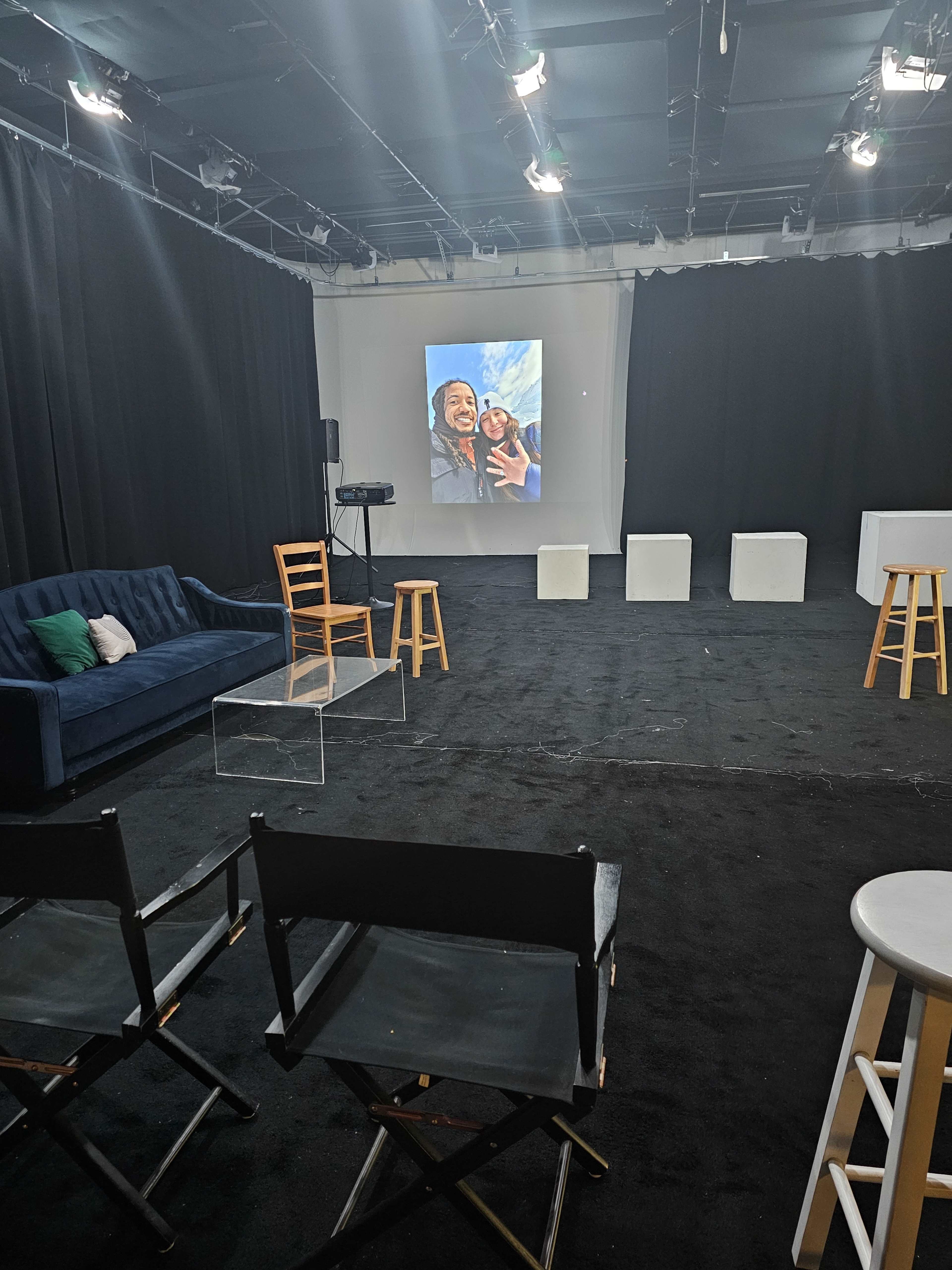 The image shows a studio space with a blue couch, a clear table, several stools, and a projector screen displaying a selfie of two people.