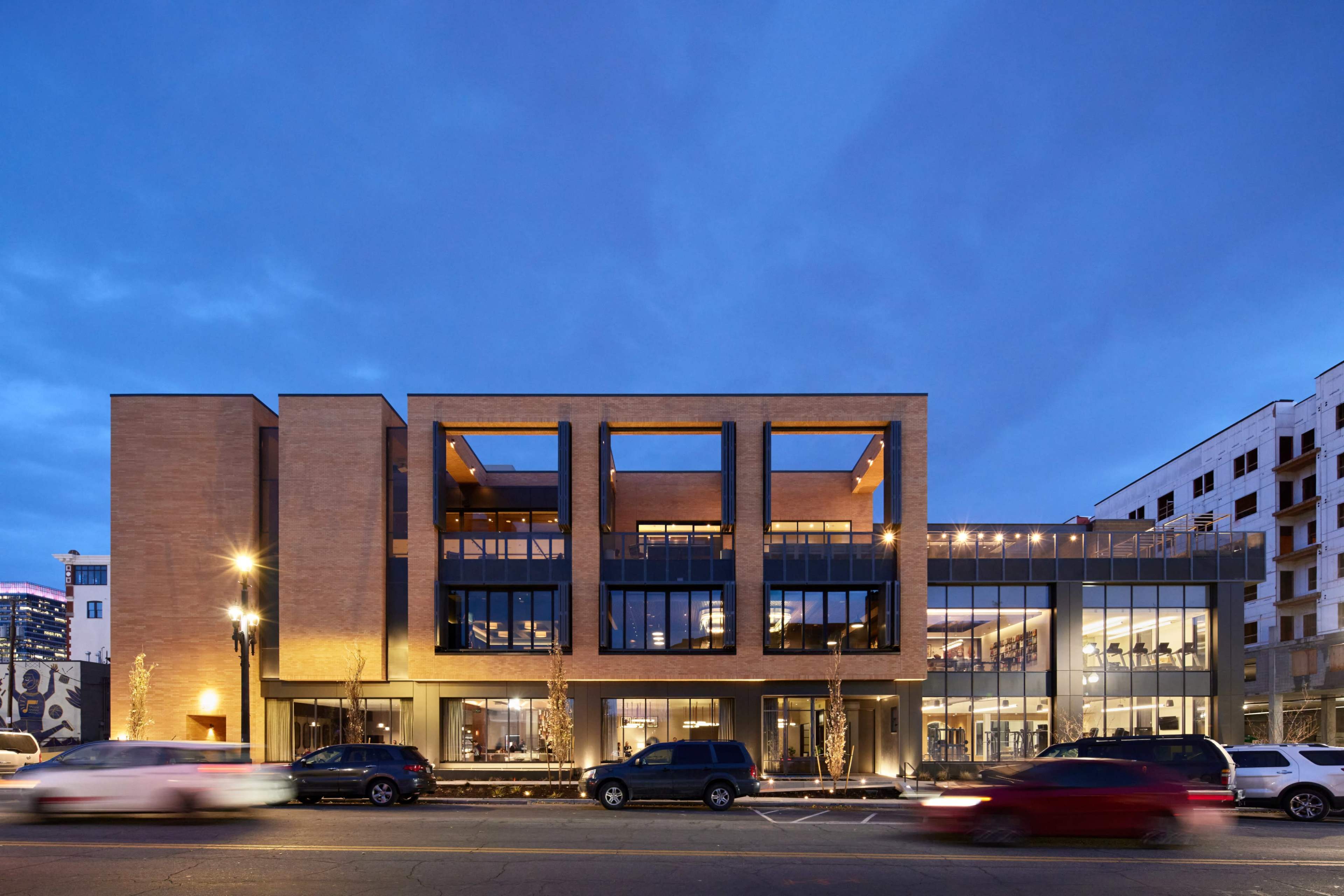 The image shows a modern multi-story building with a brick facade and large glass windows, situated along a street with cars passing by at dusk.