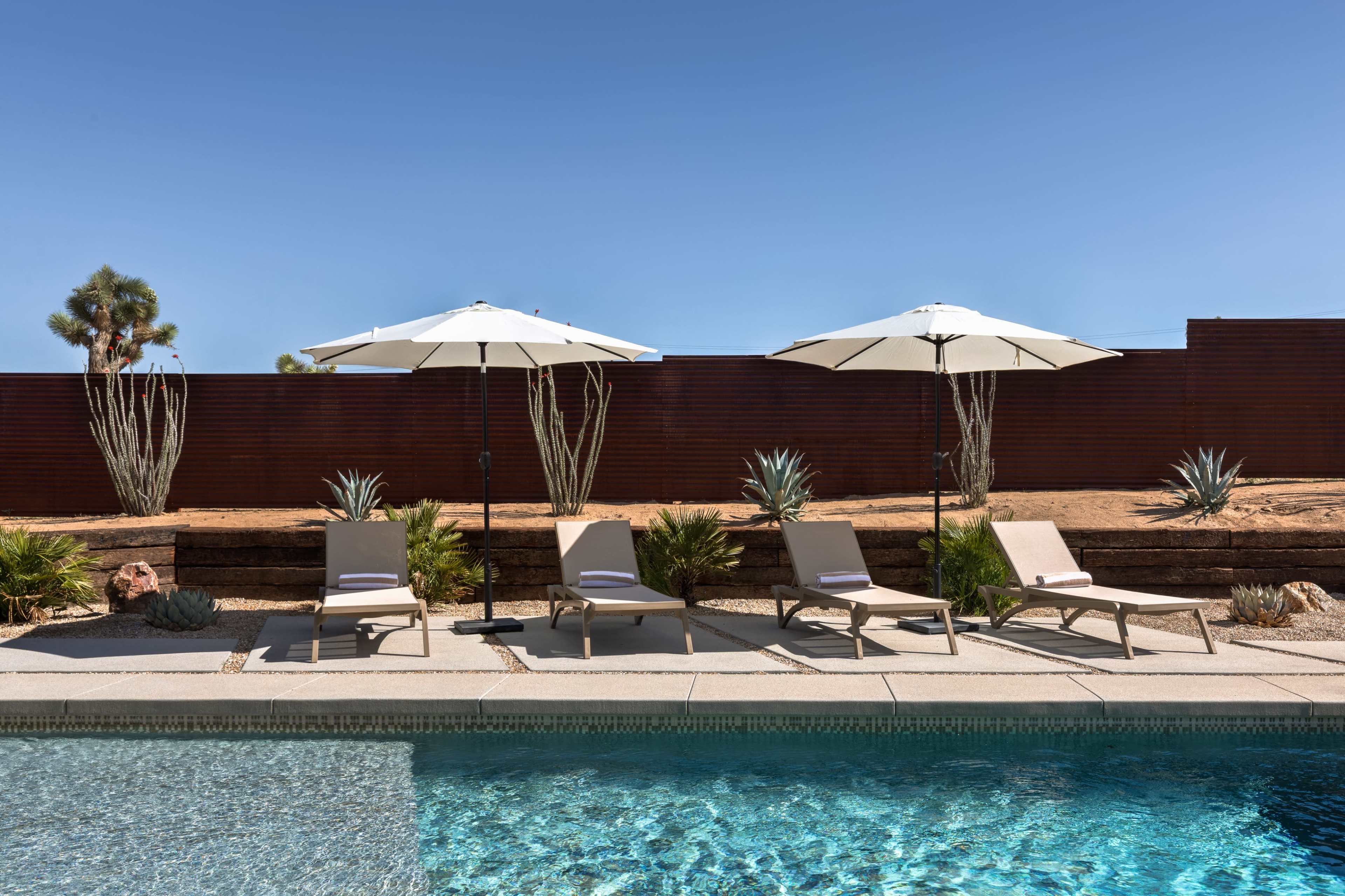 The image shows a swimming pool area with lounge chairs and white umbrellas surrounded by desert landscaping.