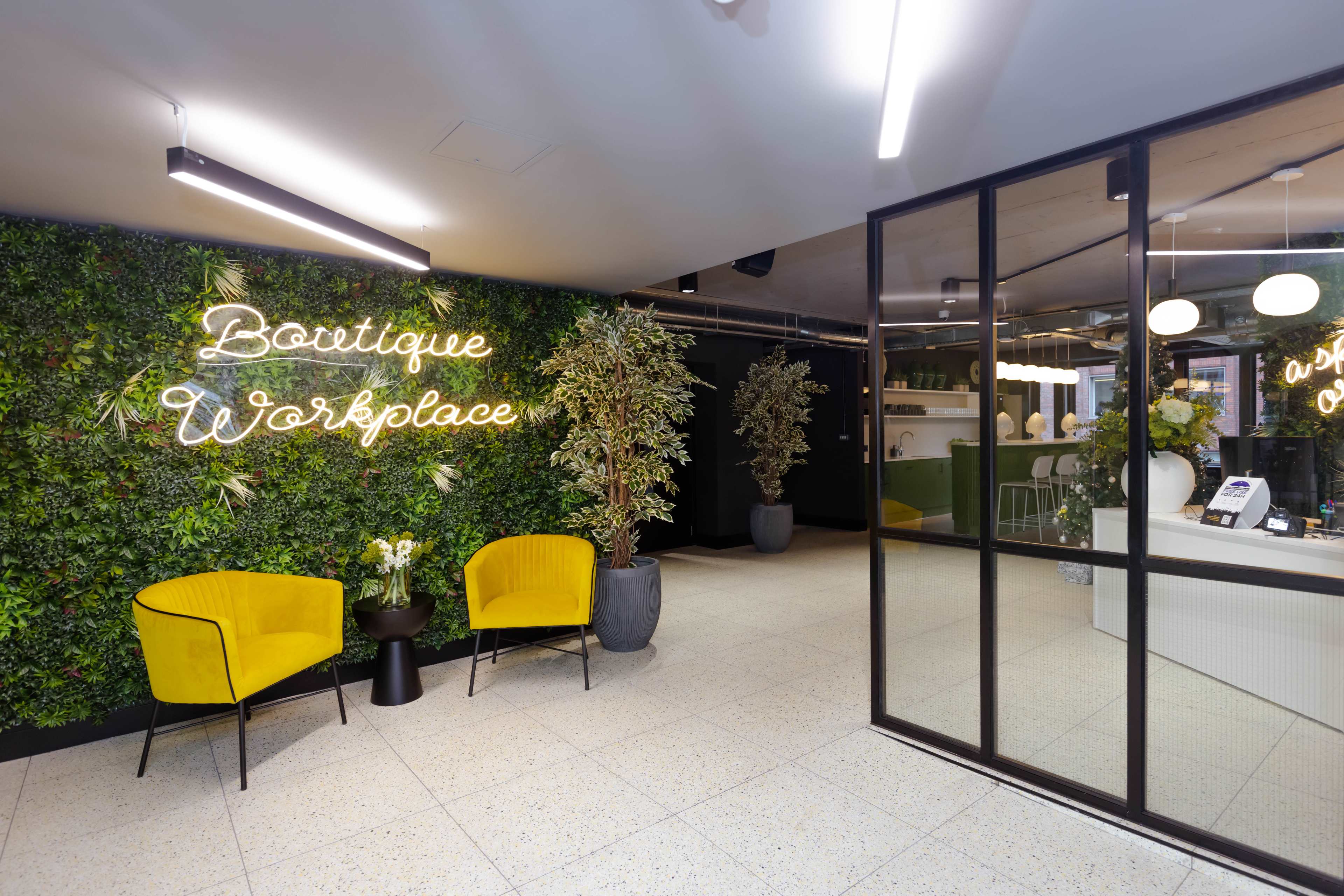 A modern workspace entrance featuring yellow chairs, greenery on the walls, and a reception area with a neon sign that reads "Boutique Workplace."