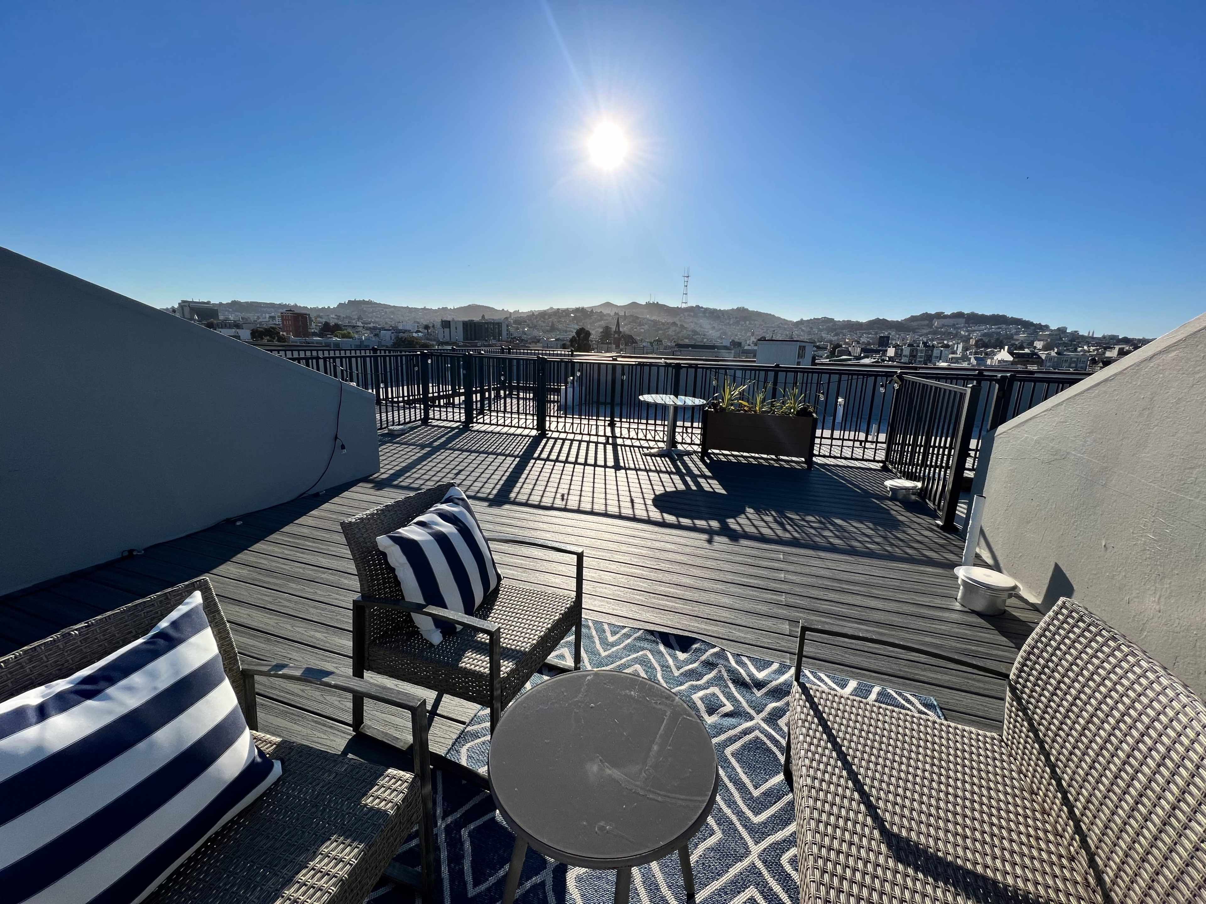 A rooftop terrace features two chairs with striped cushions, a small table, and a sun shining brightly over a cityscape.