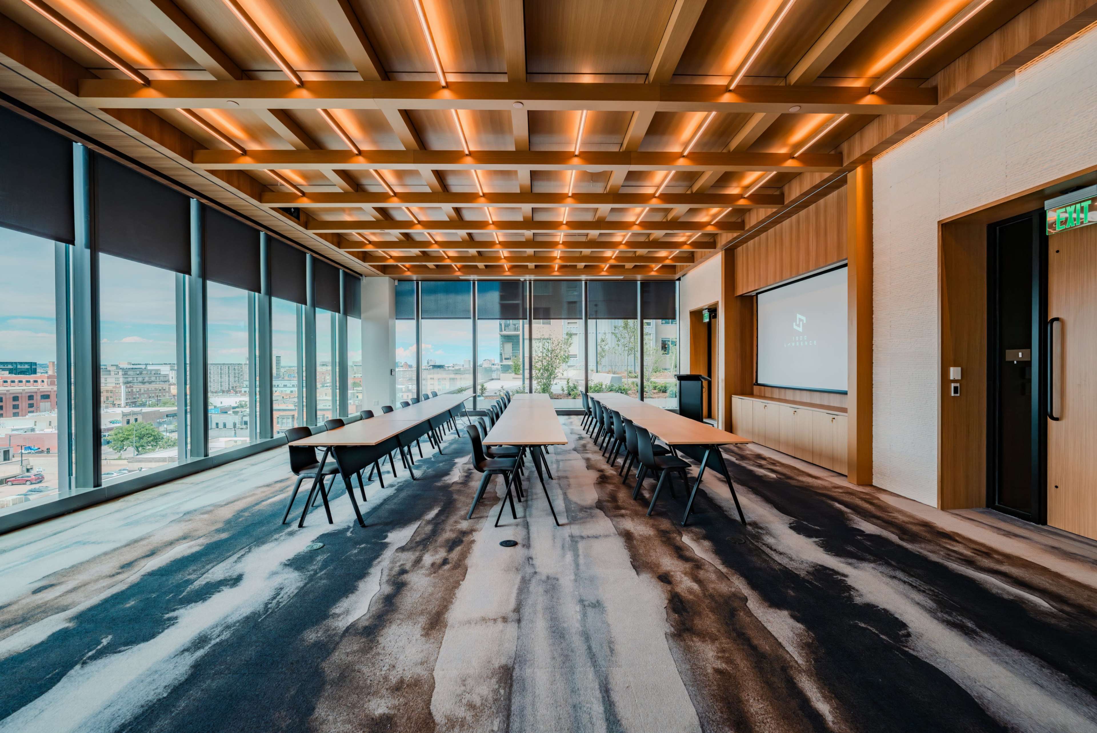 The image shows a modern meeting room with long tables arranged in a center-facing layout, large windows providing natural light, and a presentation screen on one wall.