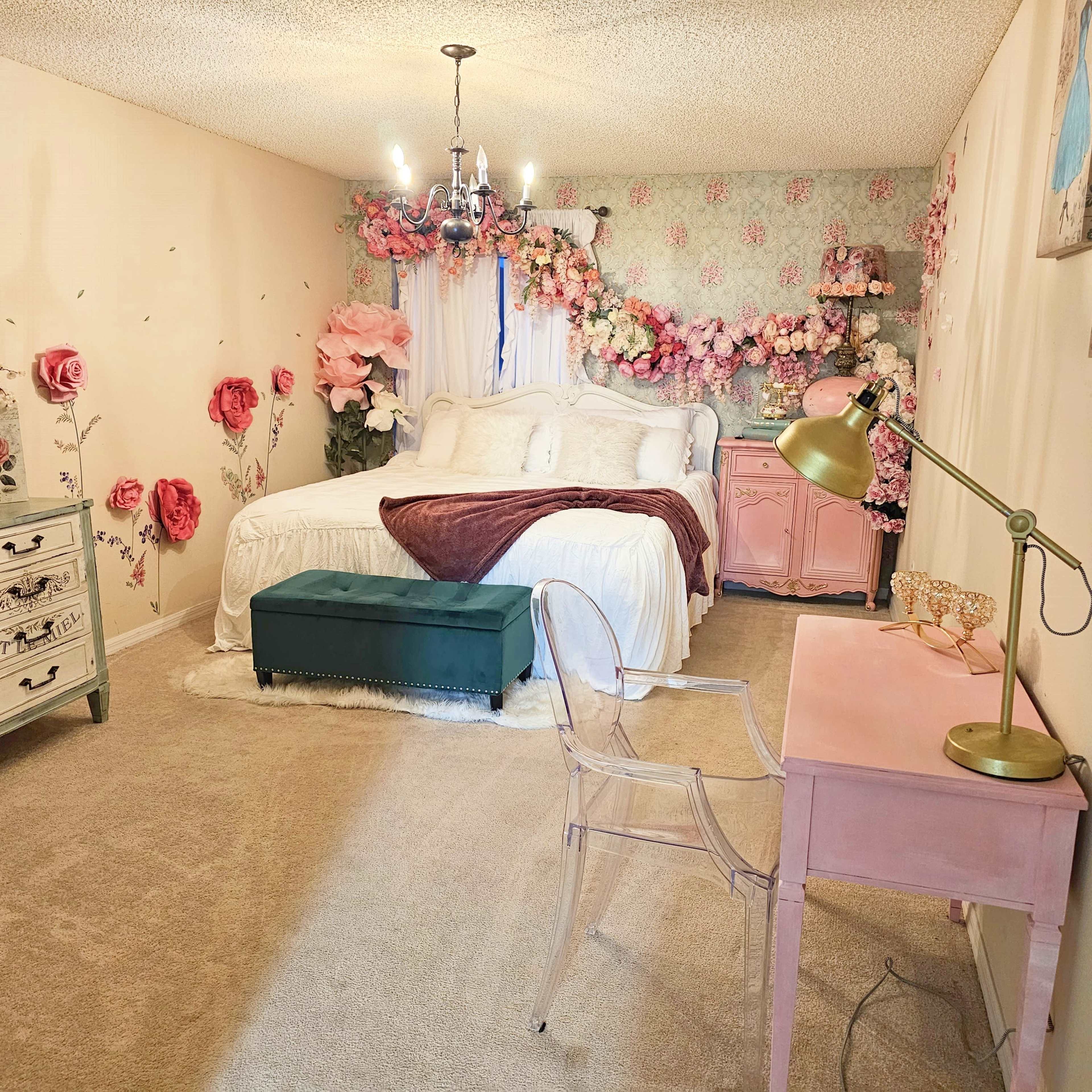 The image shows a decorated bedroom with a bed, floral accents on the walls, a small desk with a lamp, and a green ottoman.