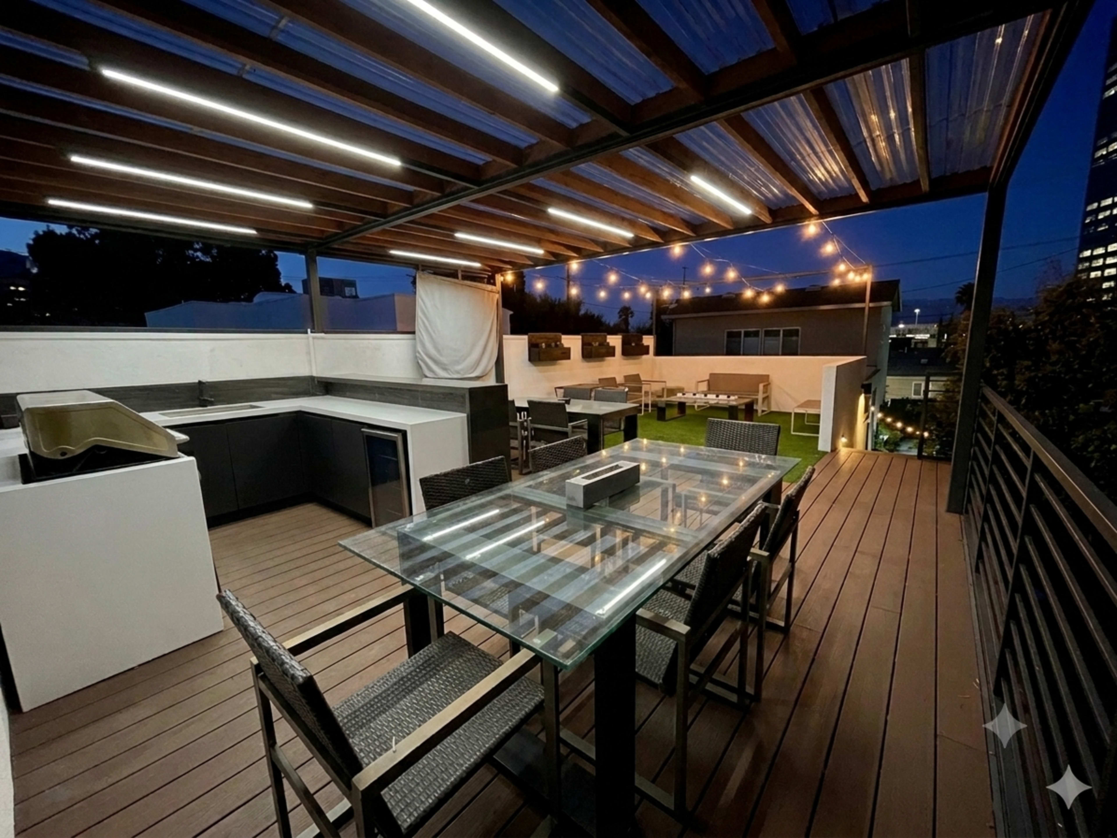 A modern rooftop terrace features a dining table with glass top, surrounded by chairs, and an outdoor kitchen area under a canopy of lights.