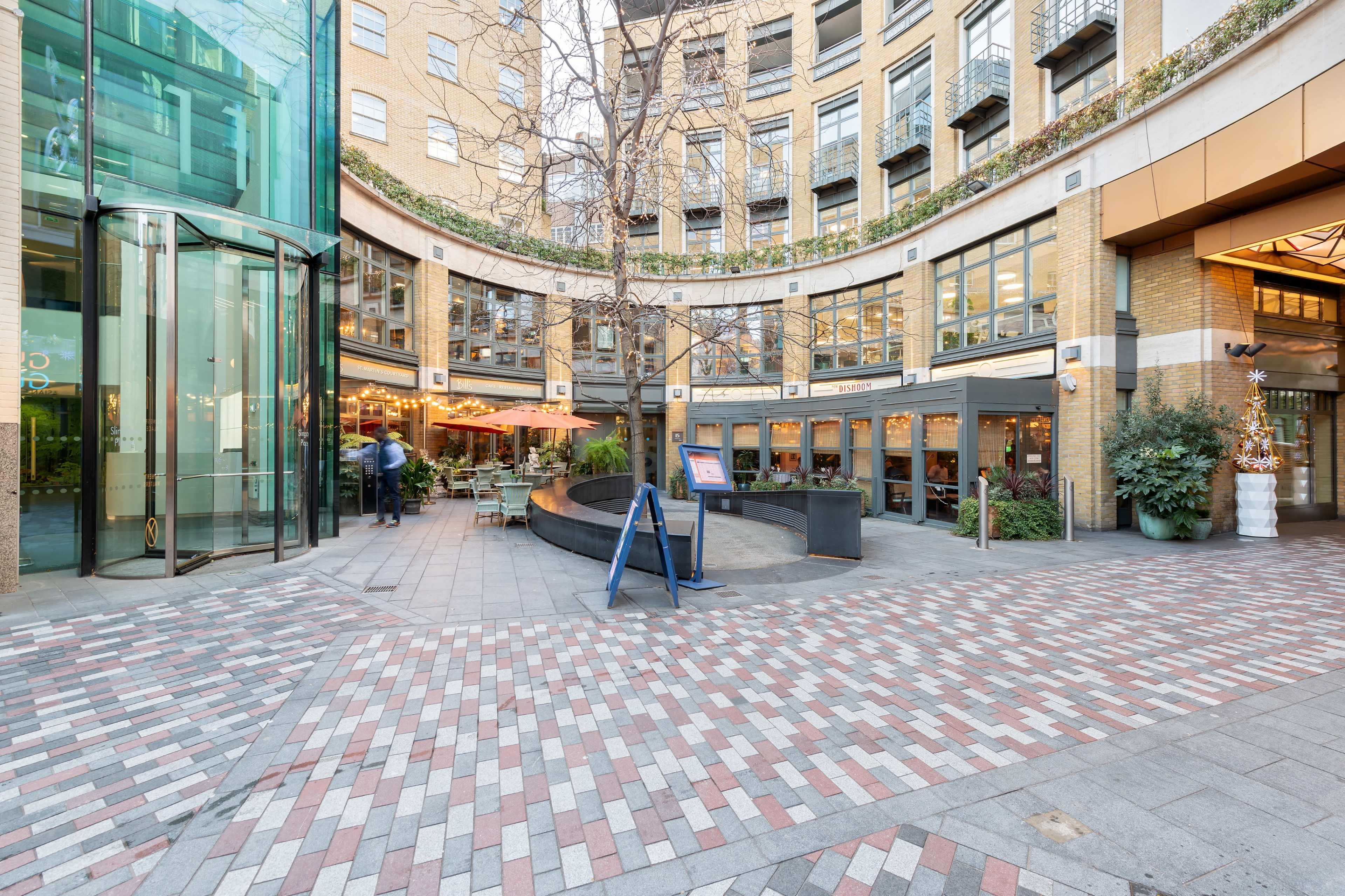 The image shows a modern courtyard with curved brick pathways, surrounded by glass buildings and outdoor seating areas at restaurants.