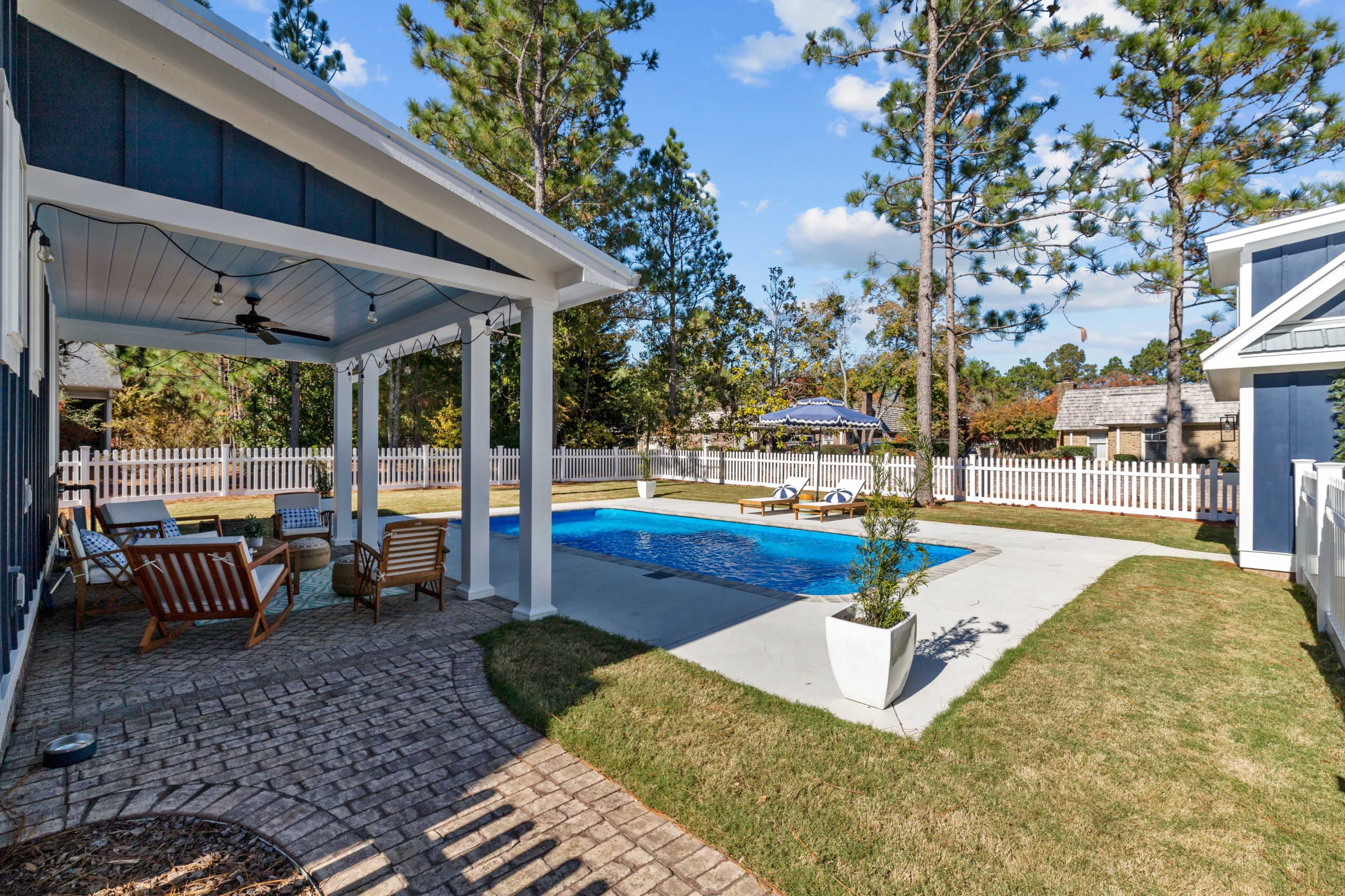 The image shows a backyard with a swimming pool surrounded by a patio area, featuring lounge chairs and chairs beneath a sheltered porch, while a white picket fence borders the space.