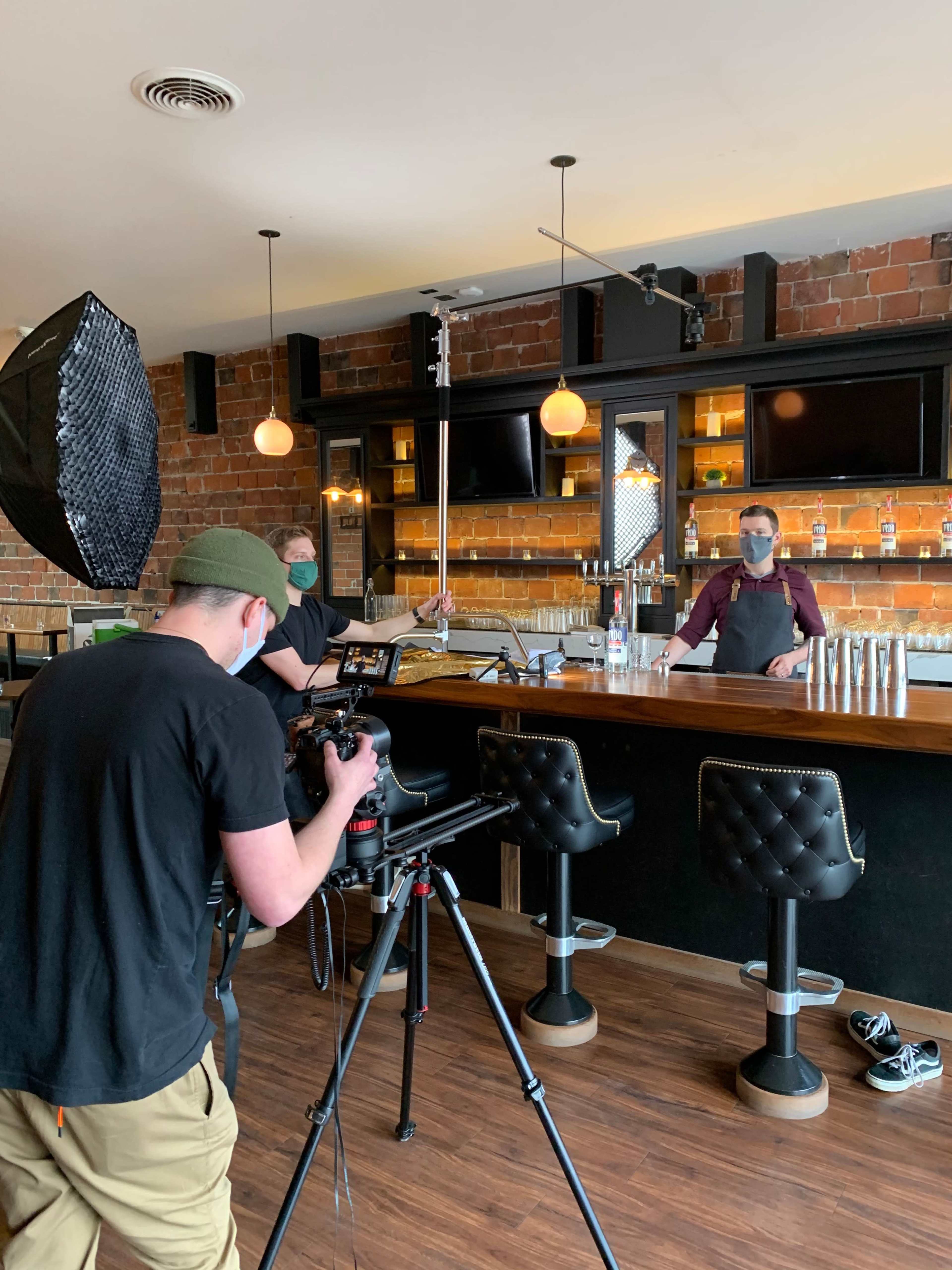 A camera operator films a bartender at a bar, while another person holds a light umbrella in the background.