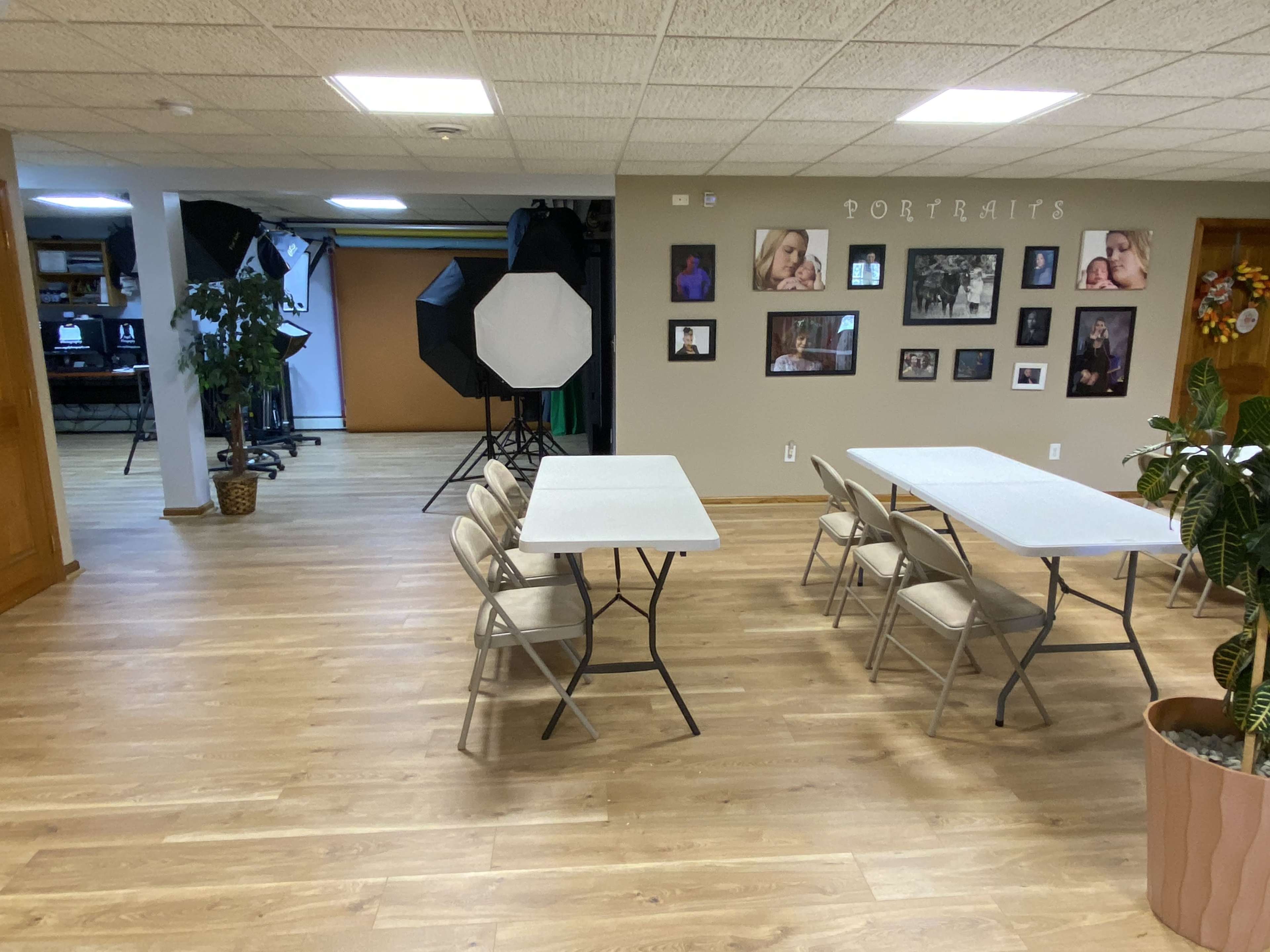 The image shows a spacious, multi-purpose room with folding tables arranged for meetings, a photography setup in the background, and various portraits displayed on the wall.