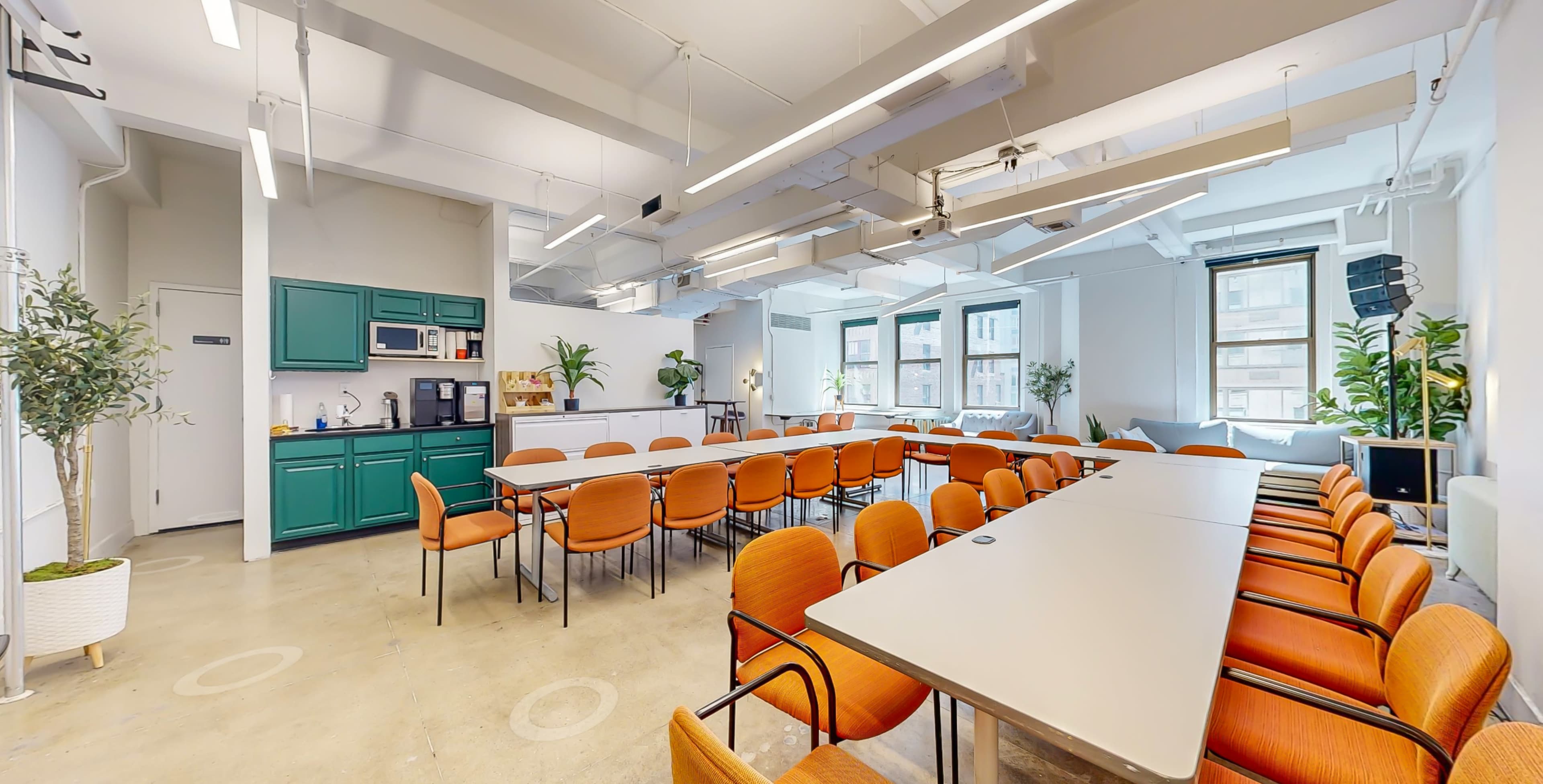 A modern conference room features long tables with orange chairs, green cabinetry, and large windows allowing natural light.