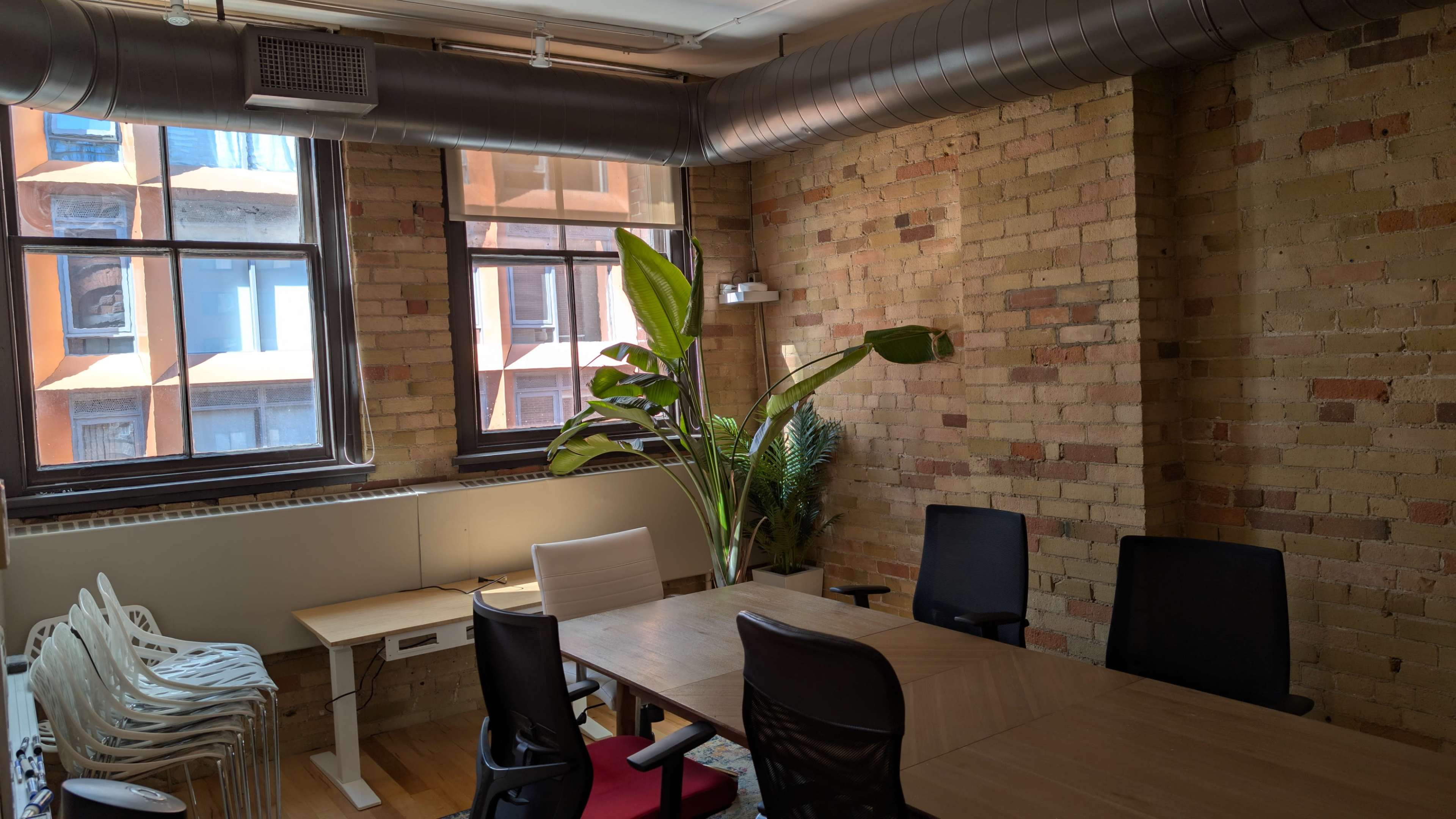 The image shows a conference room with a wooden table, several black office chairs, a potted plant, and large windows displaying a brick facade outside.