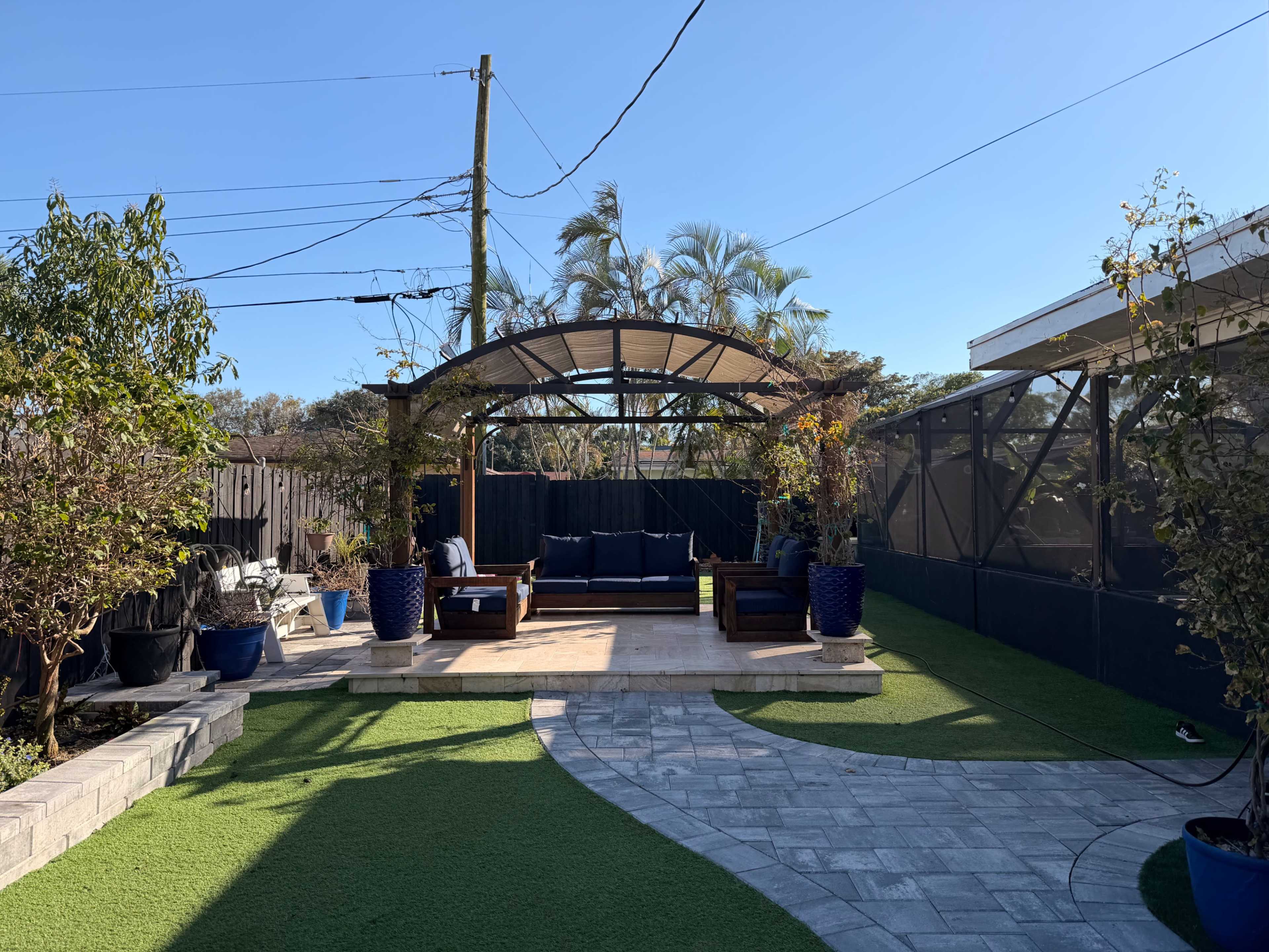 The image shows a landscaped backyard featuring a wooden pergola over a stone patio, surrounded by plants and a lawn area.