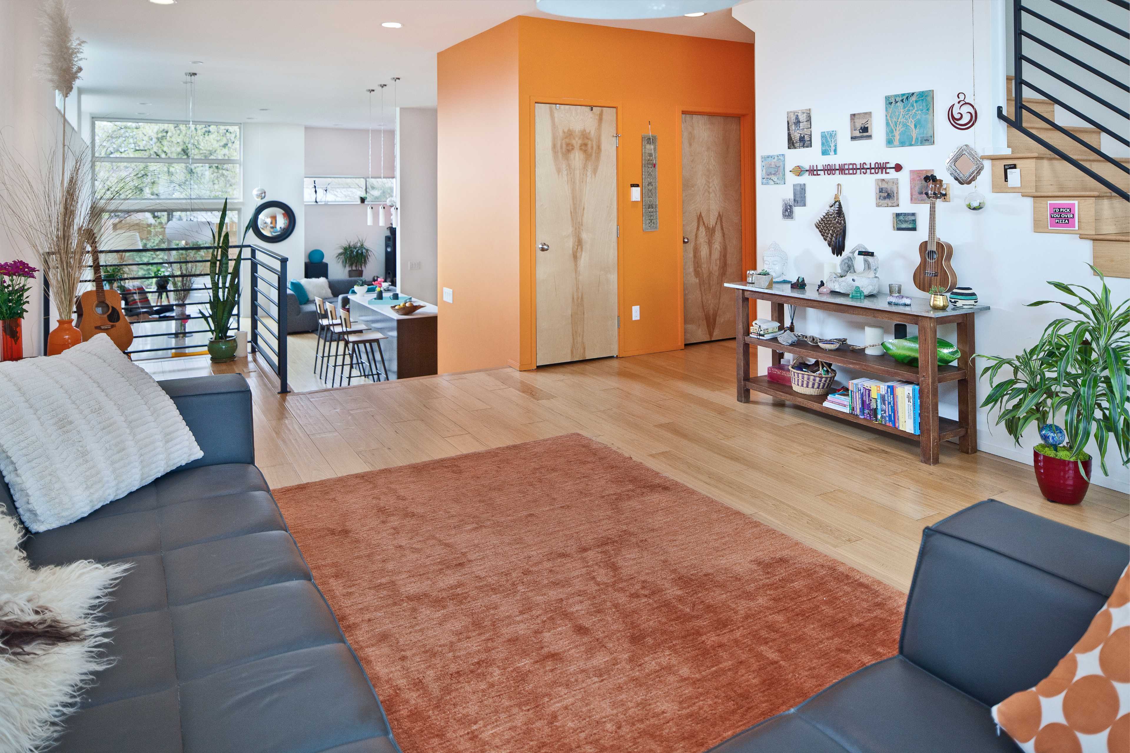 The image shows a modern living room with a large orange rug, gray sofas, decorative wall art, and a view of a dining area in the background.