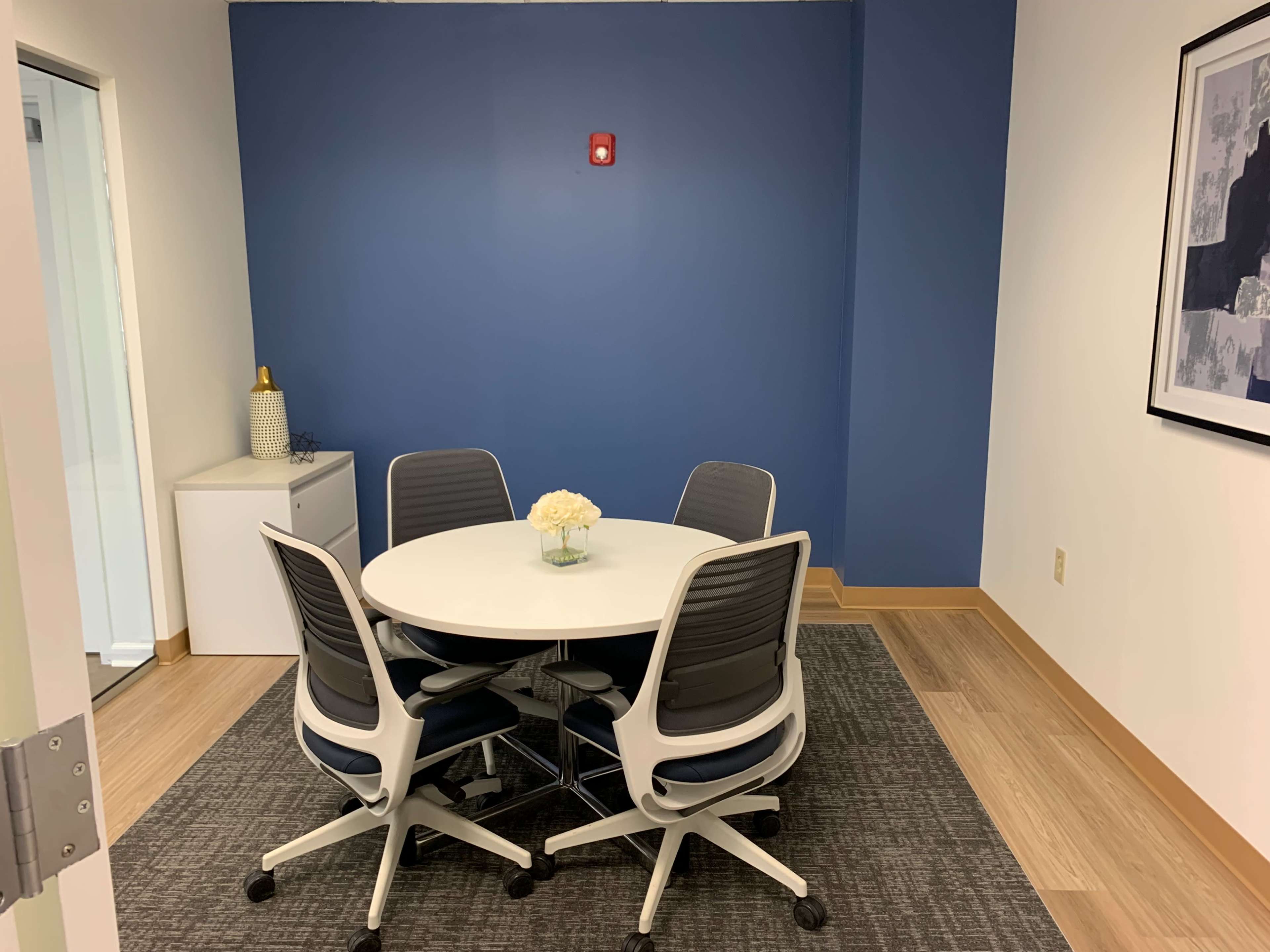 A small meeting room features a round white table surrounded by four gray chairs on a patterned rug against a blue wall.