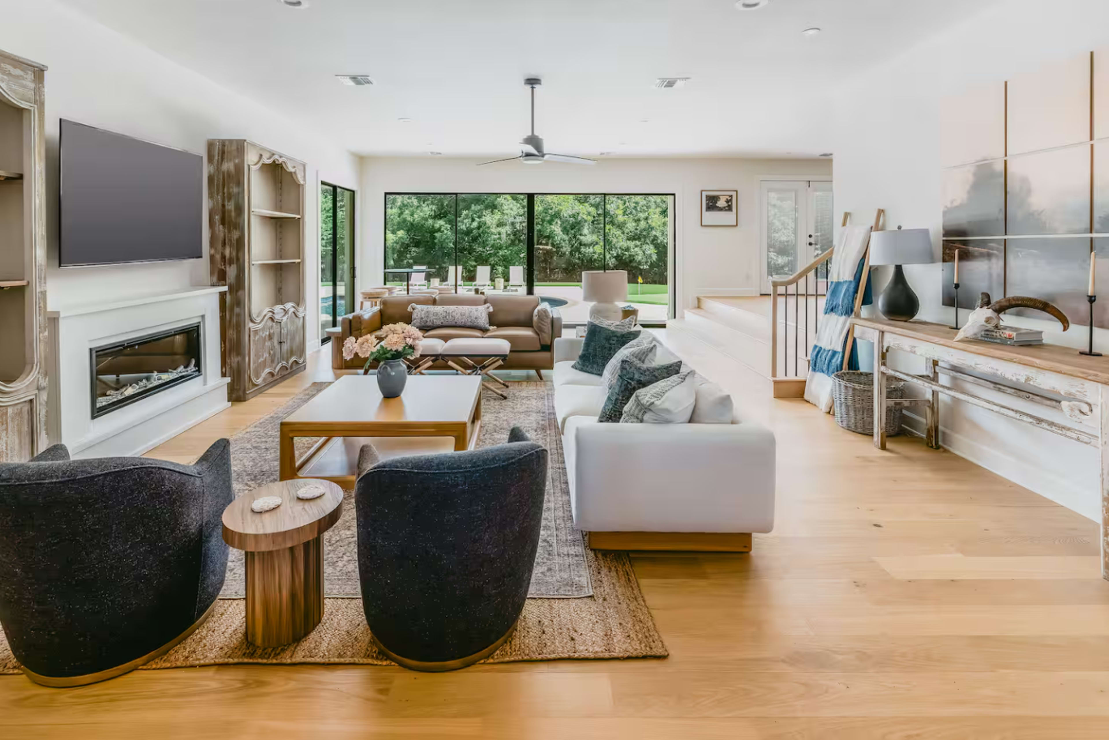 The image shows a modern living room featuring a sectional sofa, two armchairs, a coffee table, and large windows that provide a view of the outdoors.