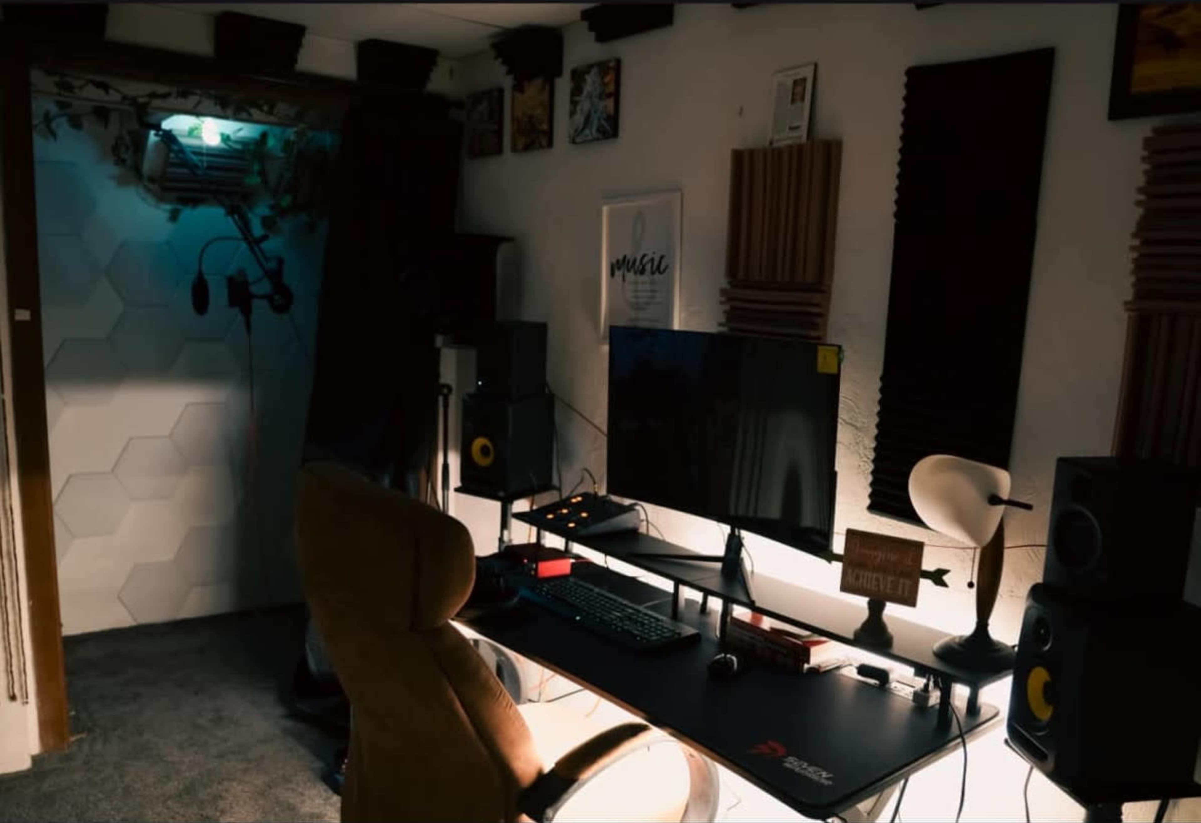 The image shows a small music studio featuring a desk with a computer monitor, speakers, and a comfortable chair, alongside soundproofing panels on the walls.