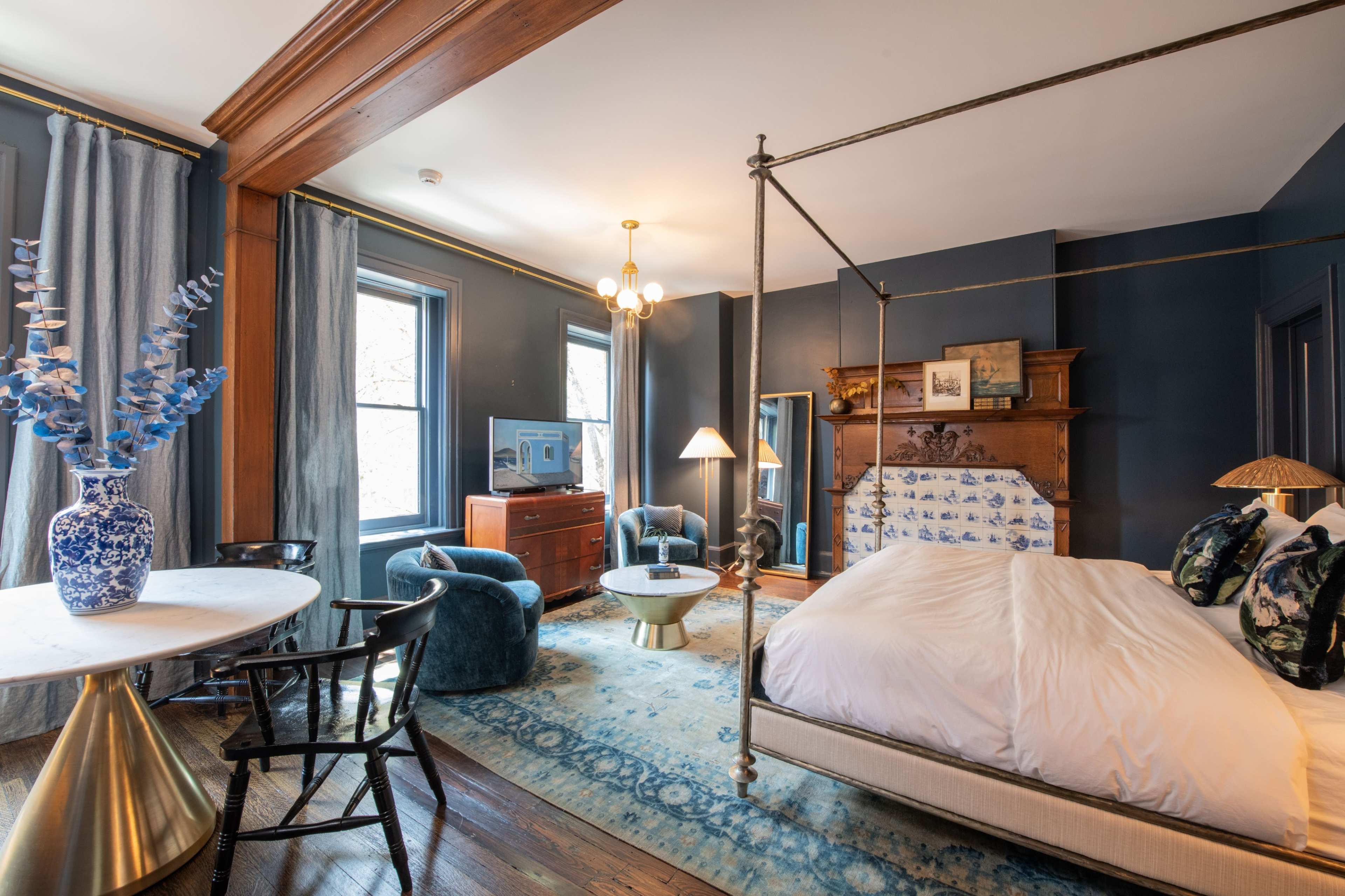 The image shows a well-furnished hotel room featuring a four-poster bed, a sitting area with blue armchairs, and a wooden dresser, all set against dark blue walls.