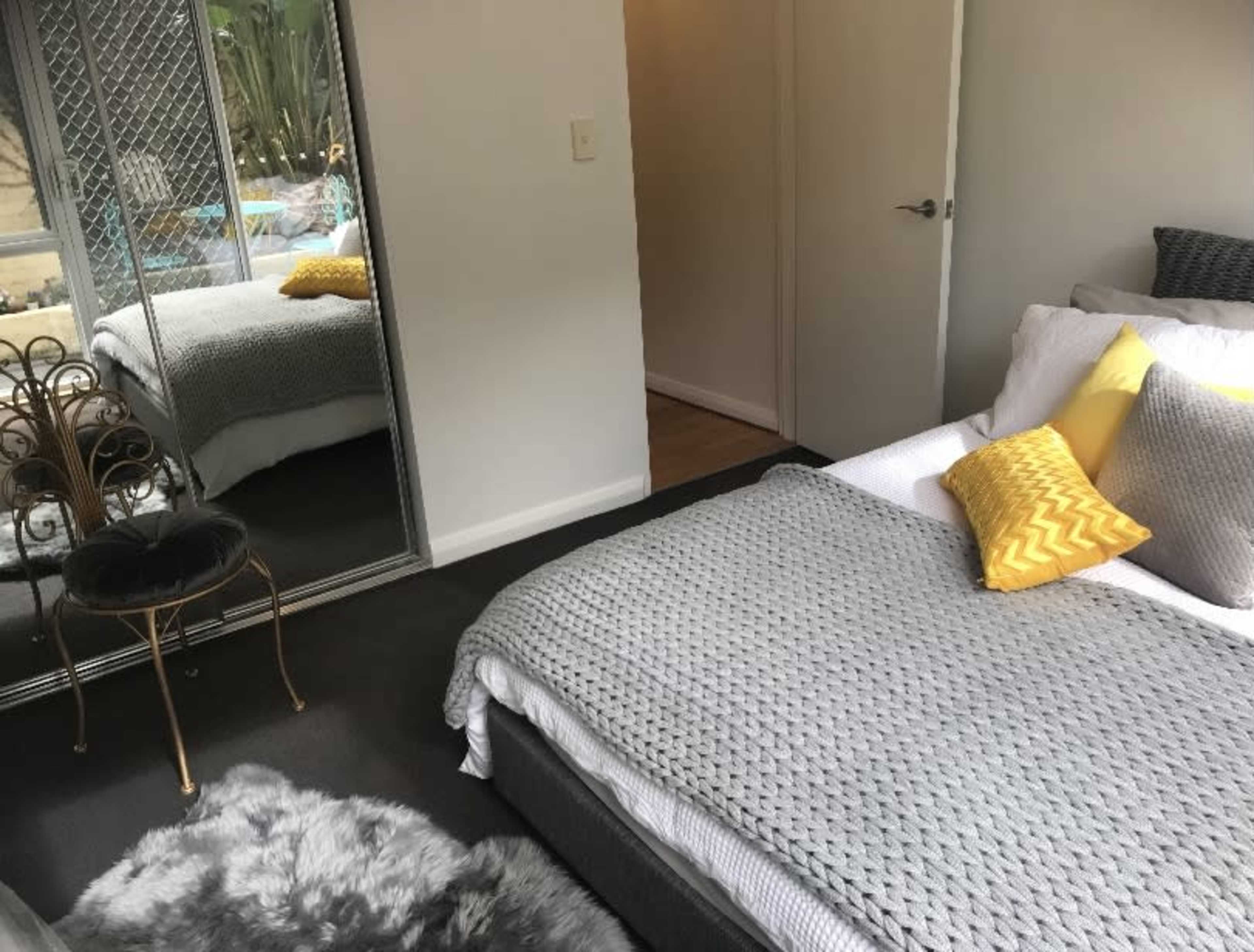 A neatly arranged bedroom features a bed with a textured gray blanket and decorative pillows, a mirrored wardrobe, and a small chair beside it.
