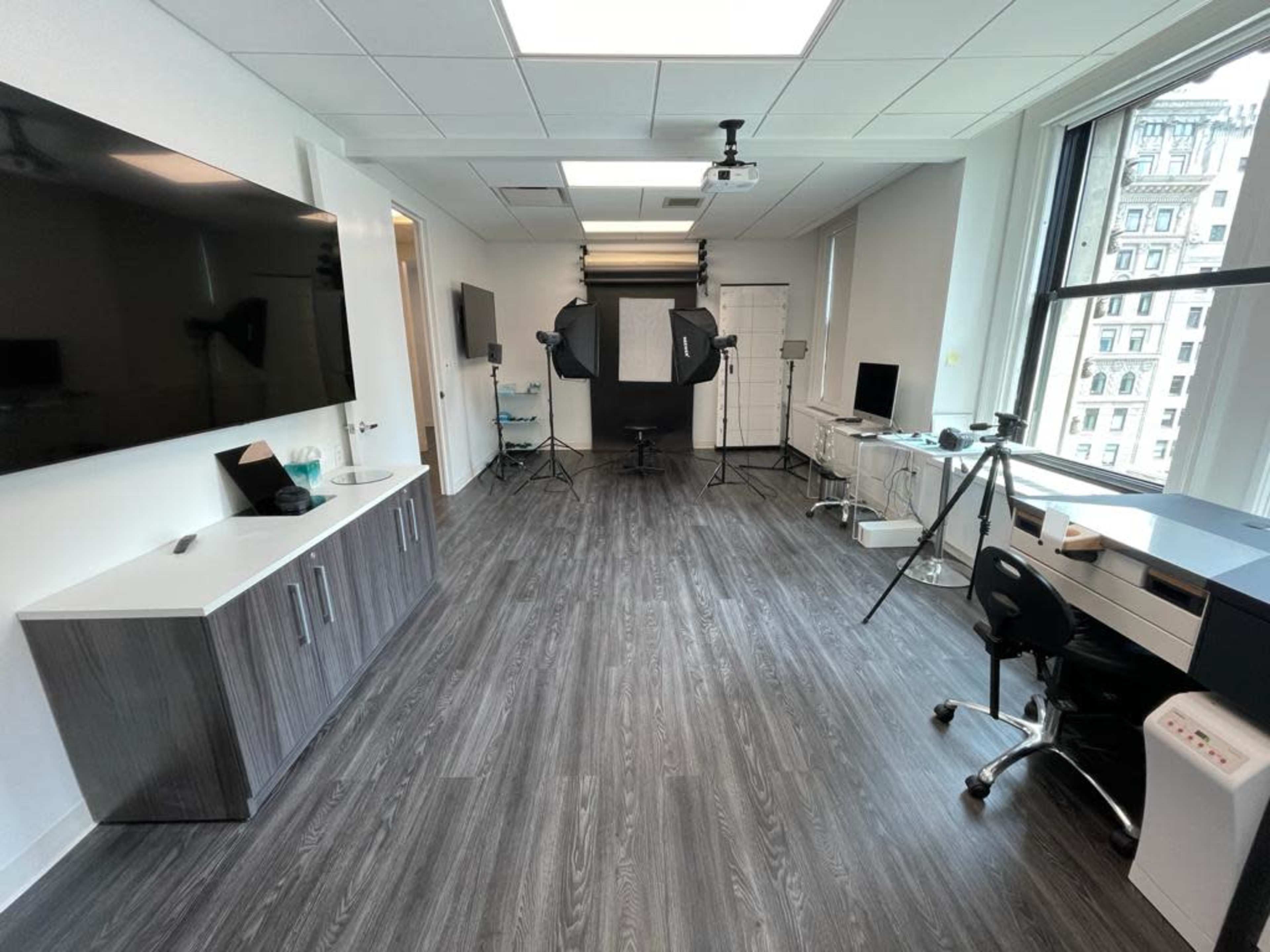 The image shows a modern studio space with a gray floor, featuring lighting equipment, a projector, and various office furniture against white walls and large windows.