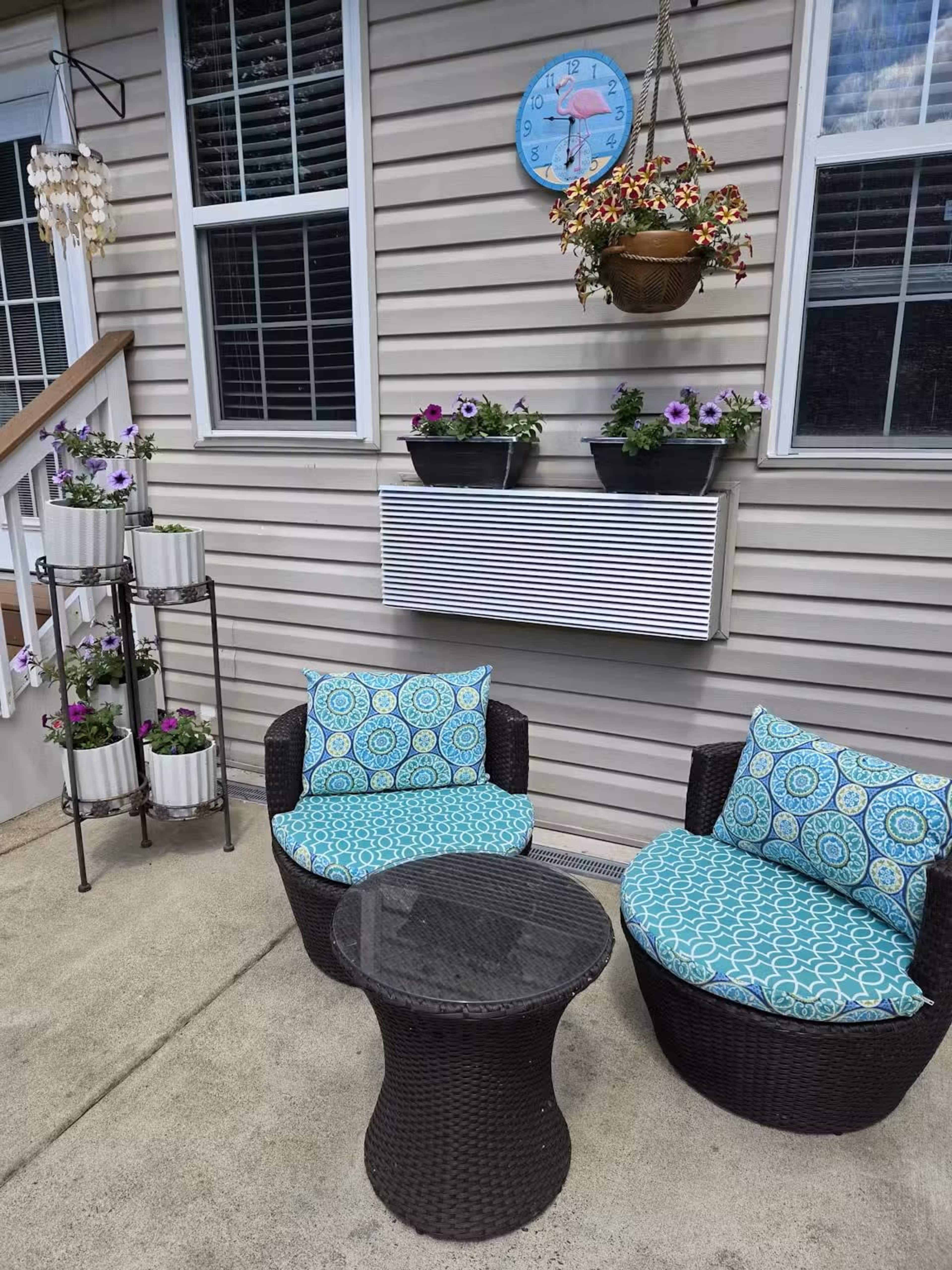 The image shows a small outdoor seating area with two round wicker chairs, a glass-topped table, a vertical plant stand, and wall-mounted flower boxes under a clock.