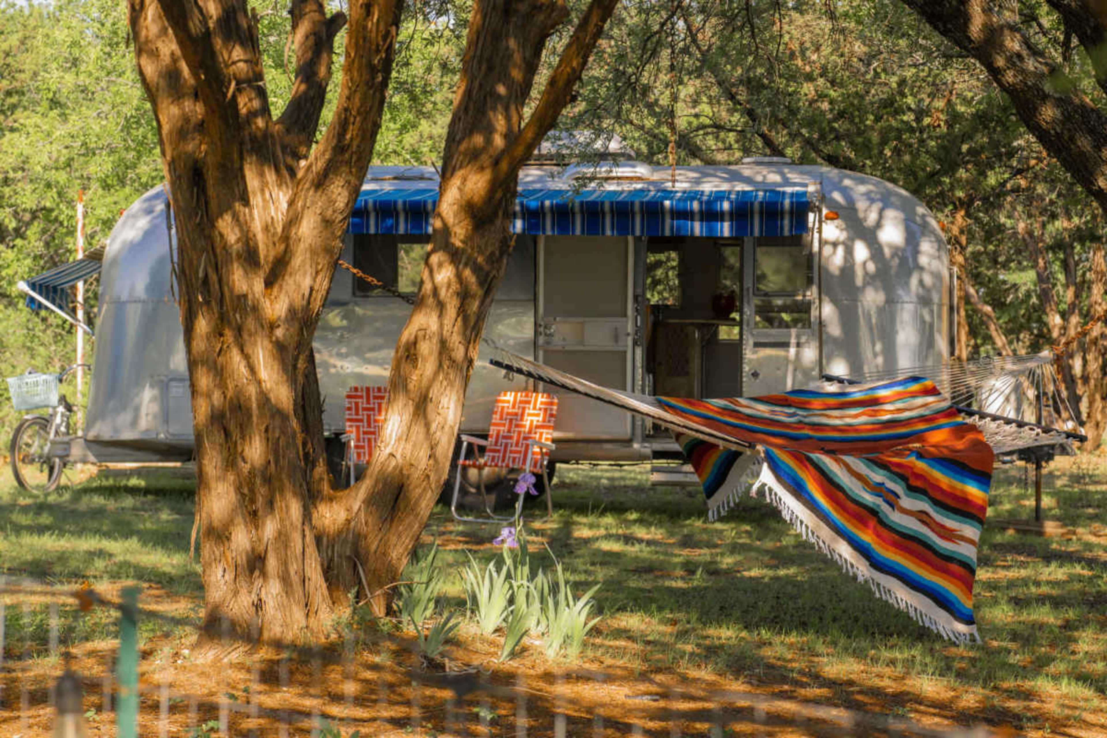 A vintage Airstream trailer is parked under trees, with a colorful blanket draped over a nearby branch and patterned chairs placed in front.