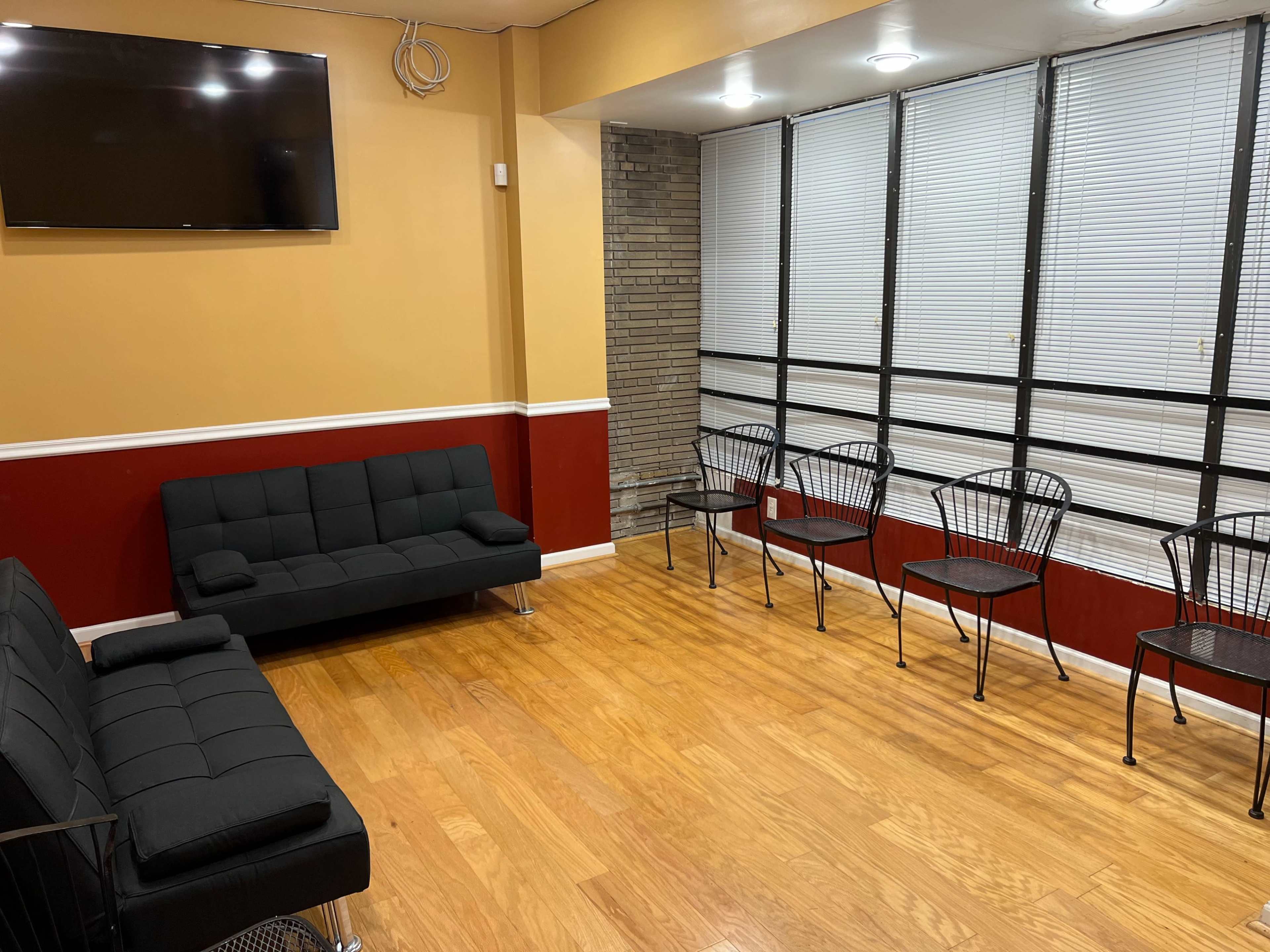 The room features a black futon, a television mounted on the wall, and several metal chairs arranged along a windowed wall.