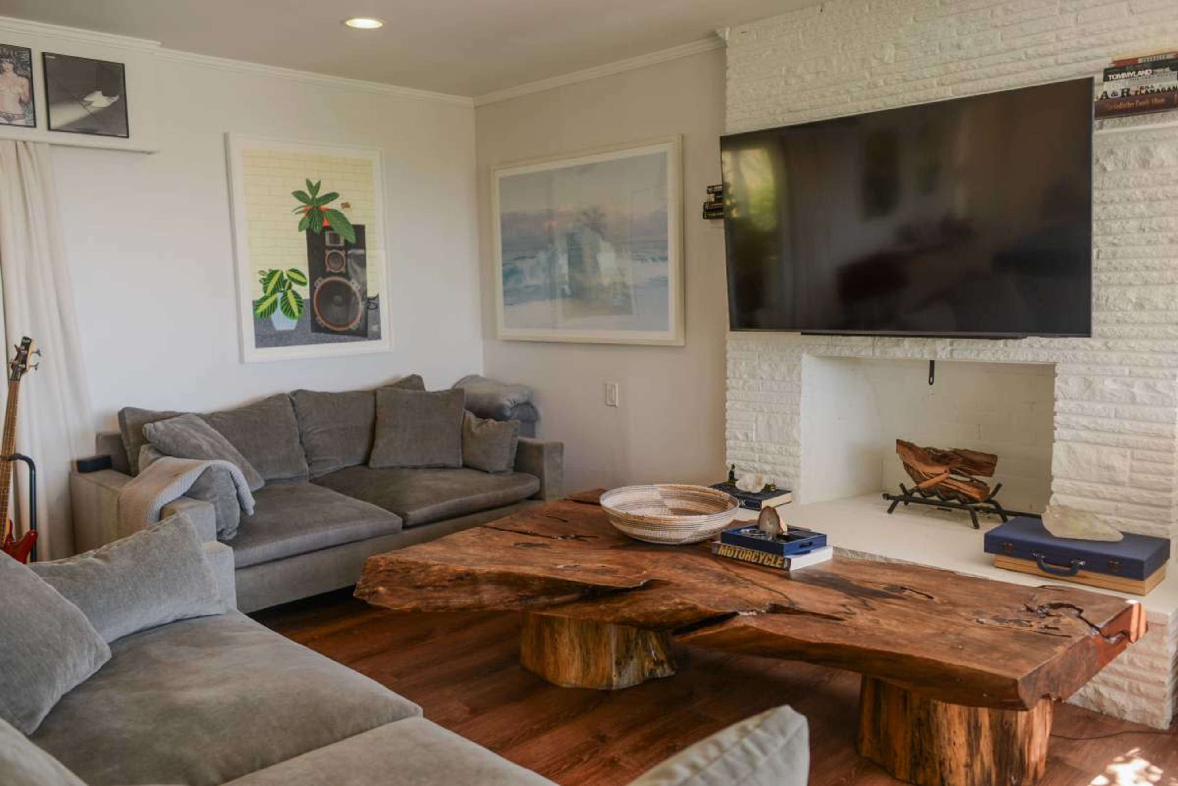 The image shows a modern living room featuring a large wooden coffee table, gray sofas, and a flat-screen TV mounted on a white brick wall.