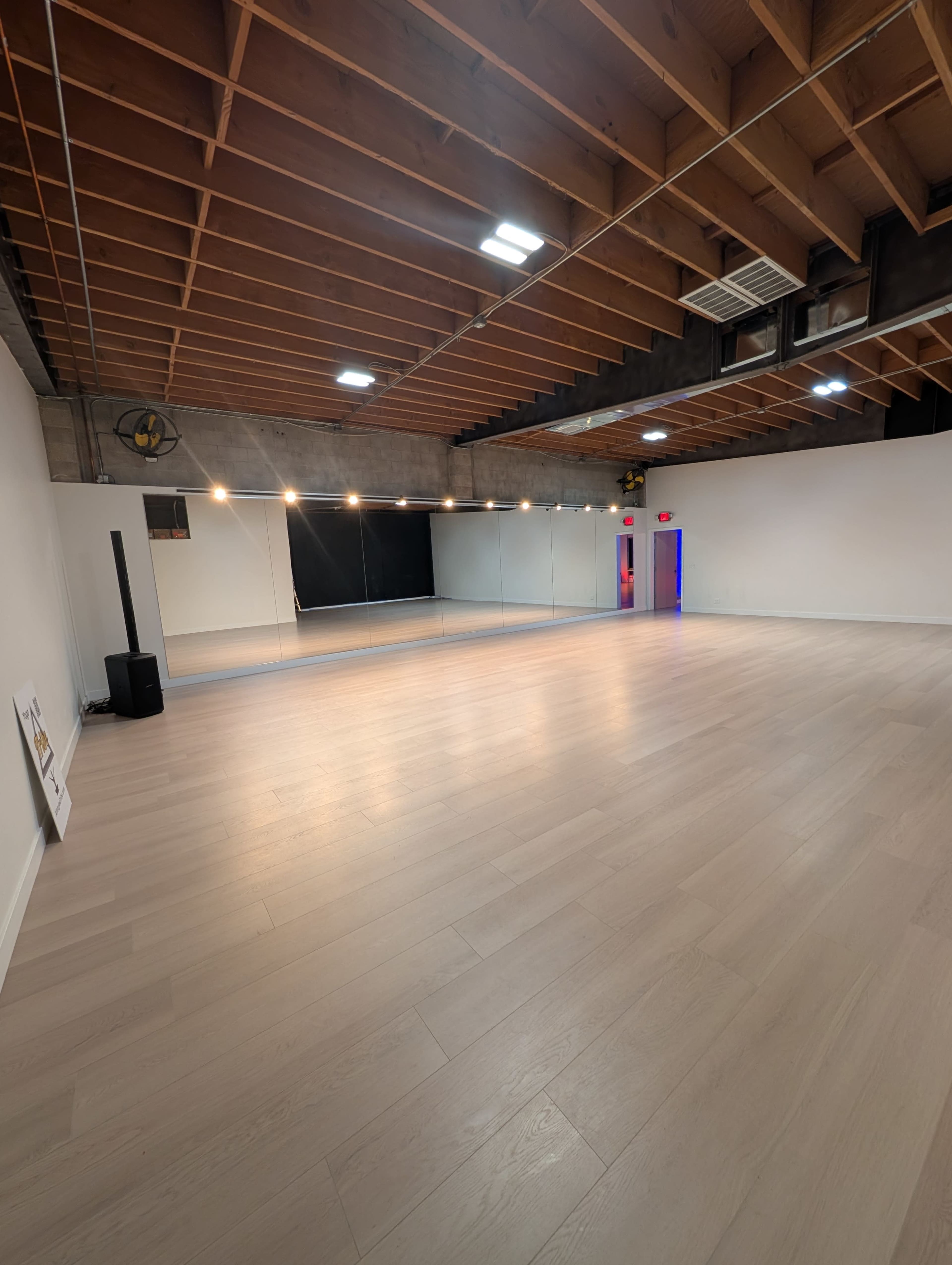The image shows a spacious, empty dance studio with wooden flooring, a large mirrored wall, and exposed ceiling beams.