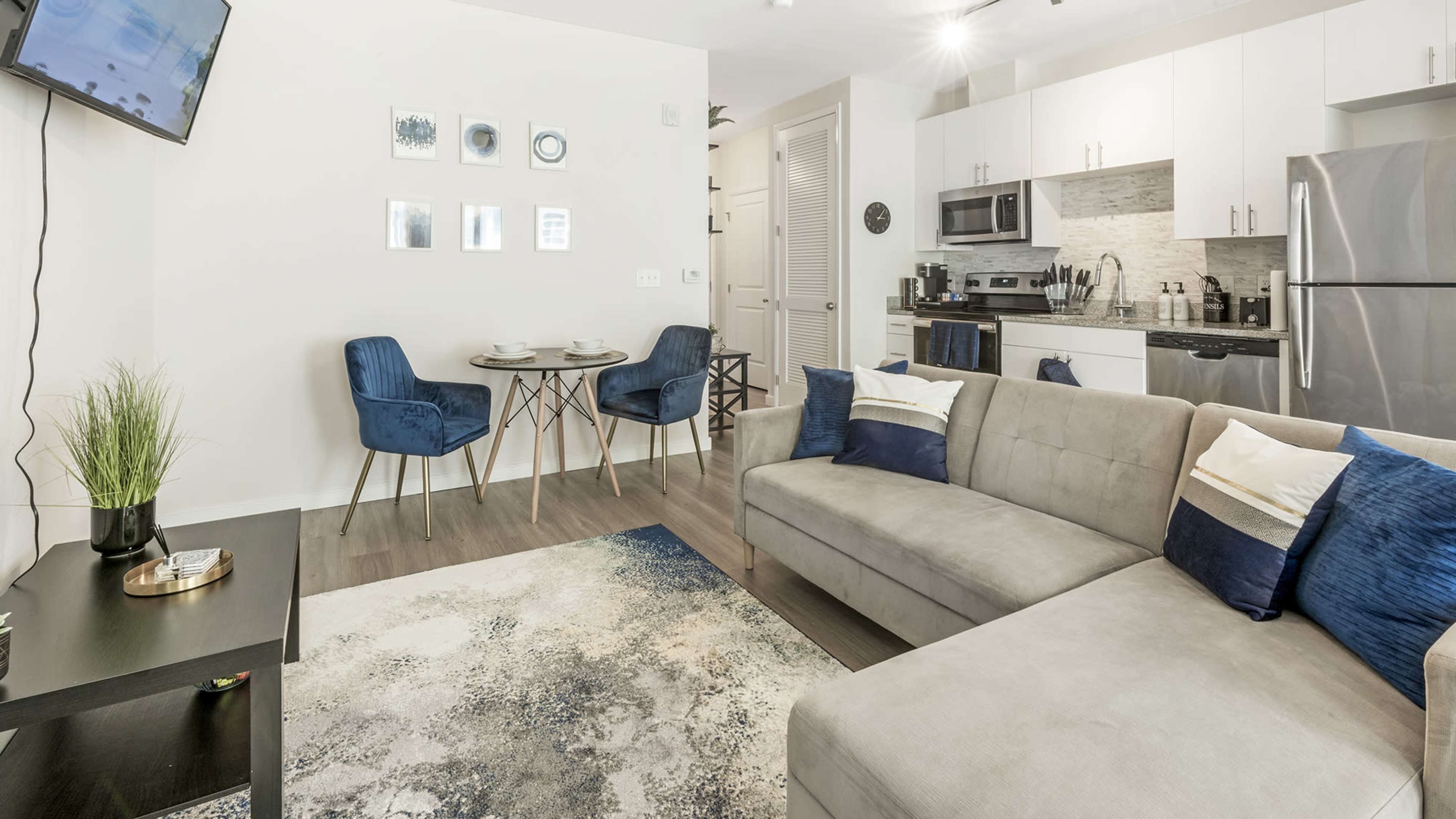 A modern living area features a gray sofa, blue accent chairs, a small dining set, and a kitchen with stainless steel appliances.