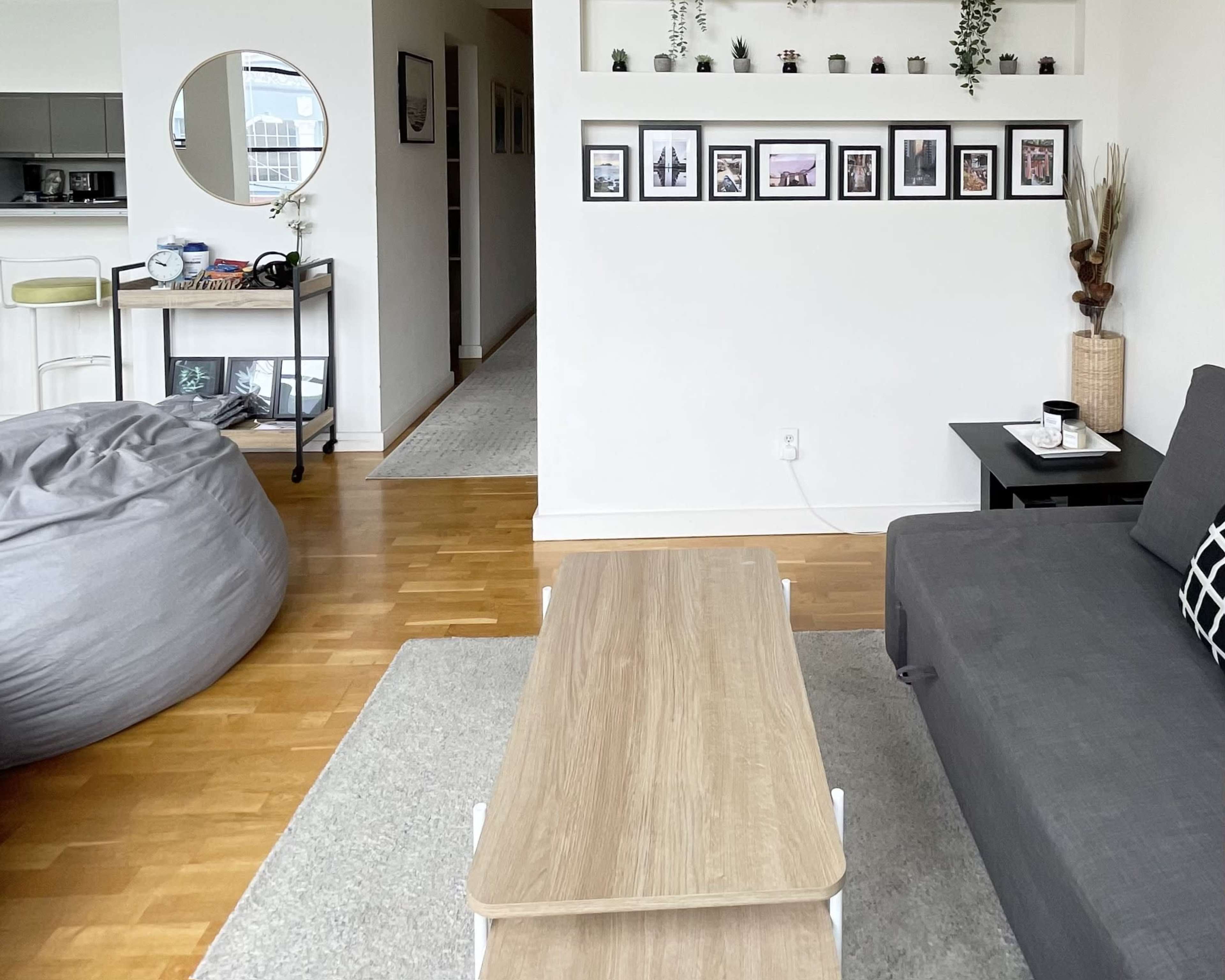 The room features a gray sofa, a wooden coffee table, and a wall adorned with framed photos, alongside decorative plants.
