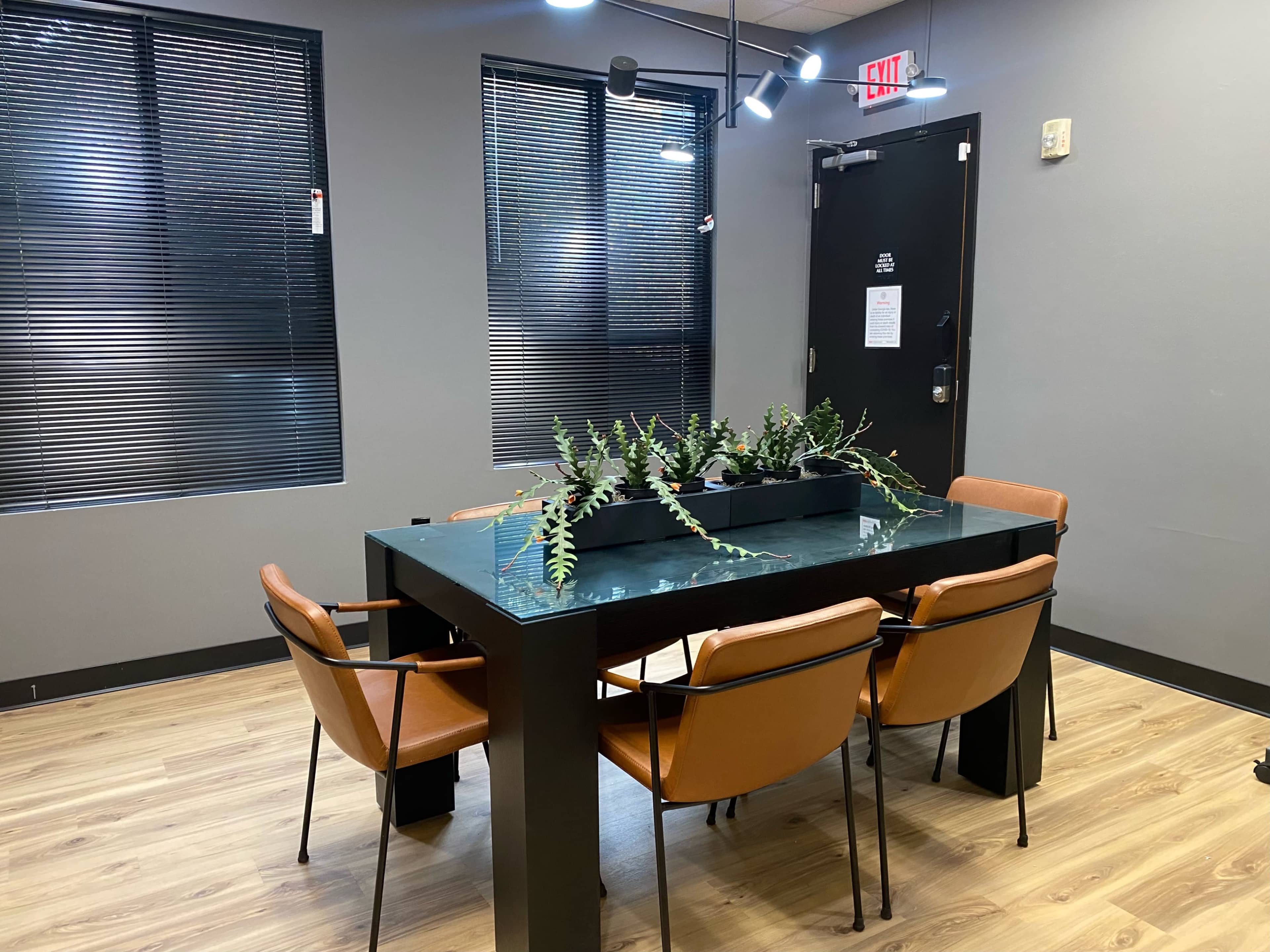 A rectangular table with a planter in the center is surrounded by four brown chairs in a well-lit room with black blinds and gray walls.