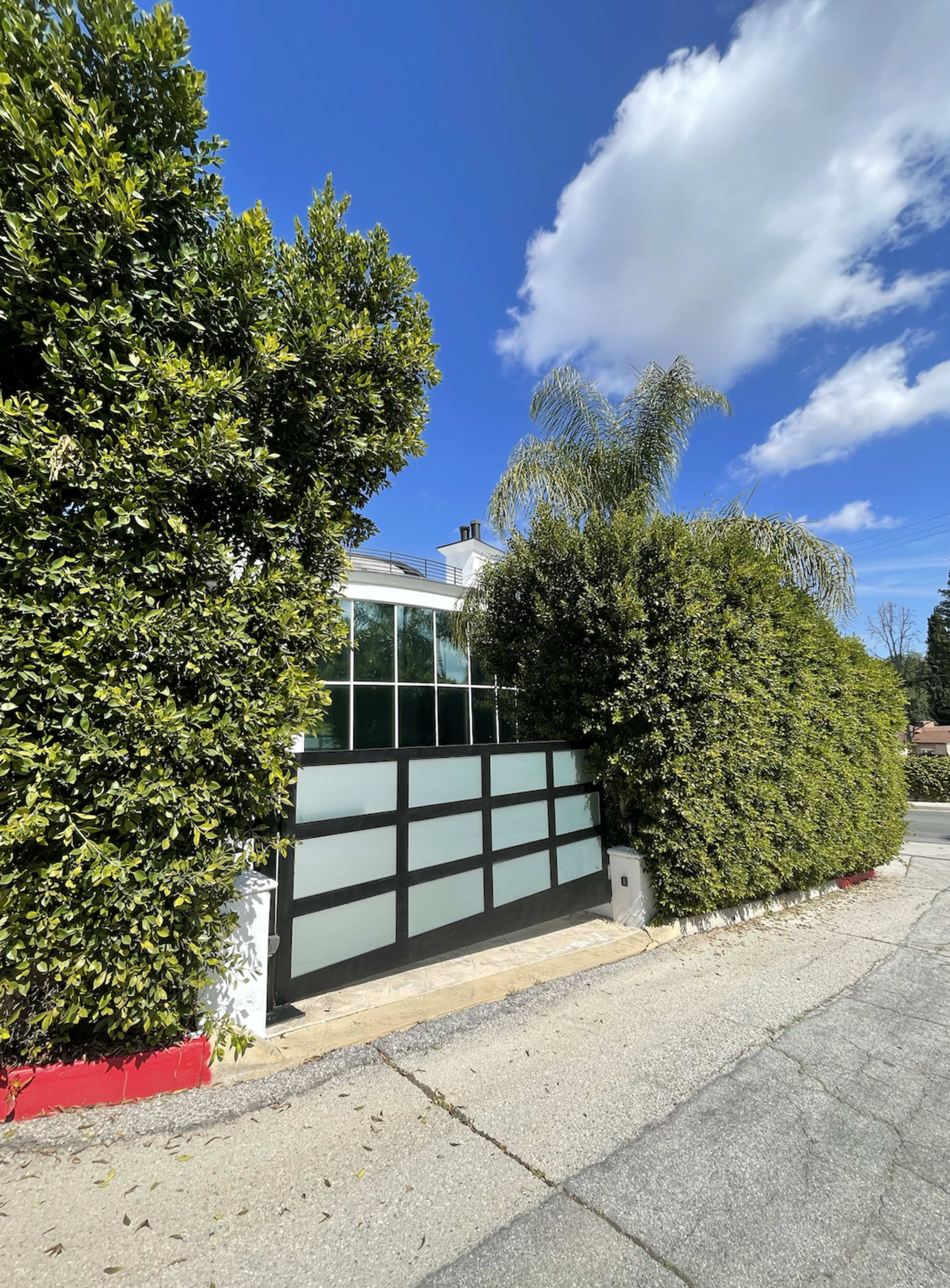 A modern gate is surrounded by tall green hedges and palm trees under a clear blue sky.