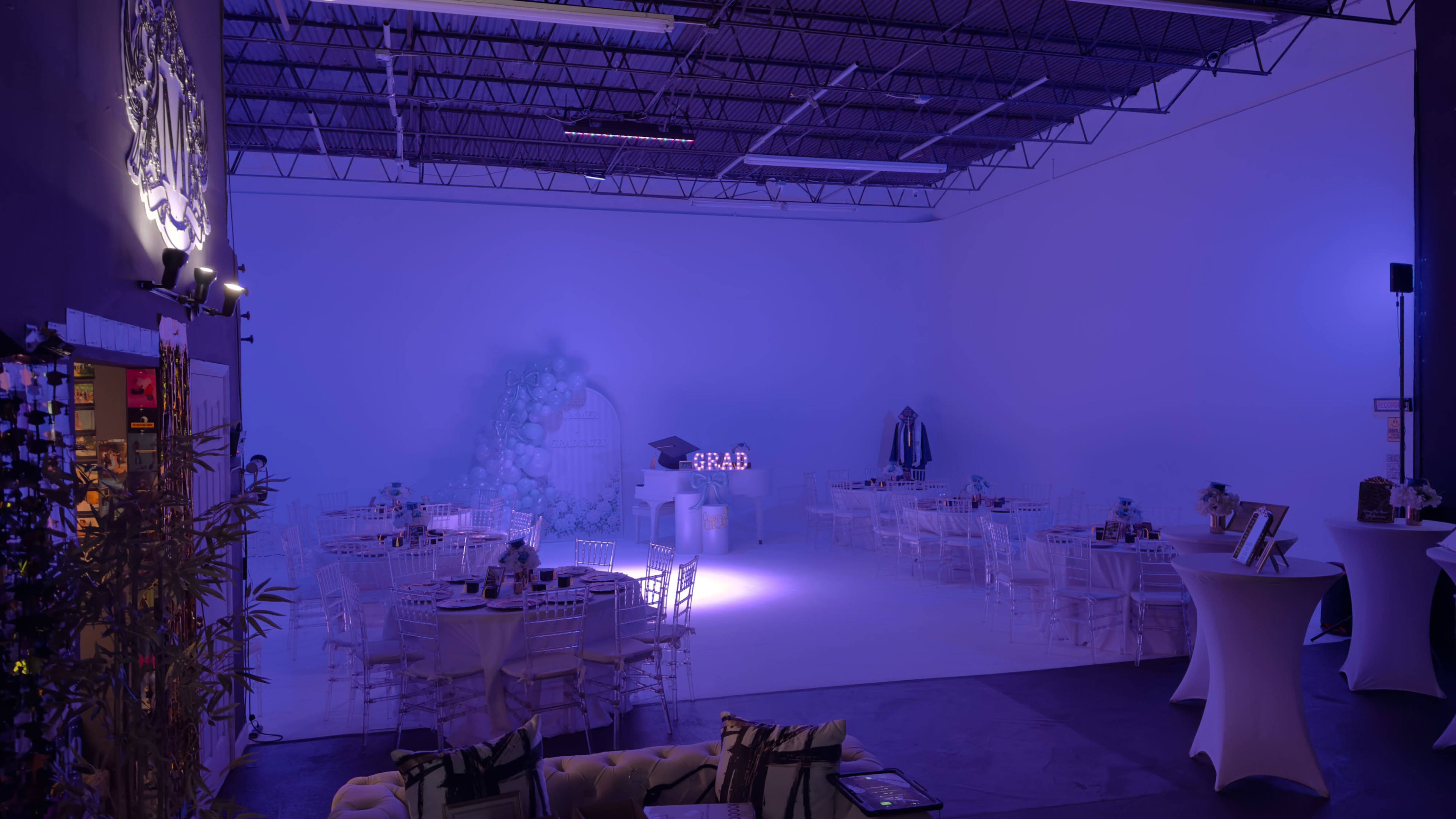 The image shows a spacious event venue set up for a gathering, featuring several round tables with white tablecloths, illuminated by soft purple lighting, and decorated with floral elements.
