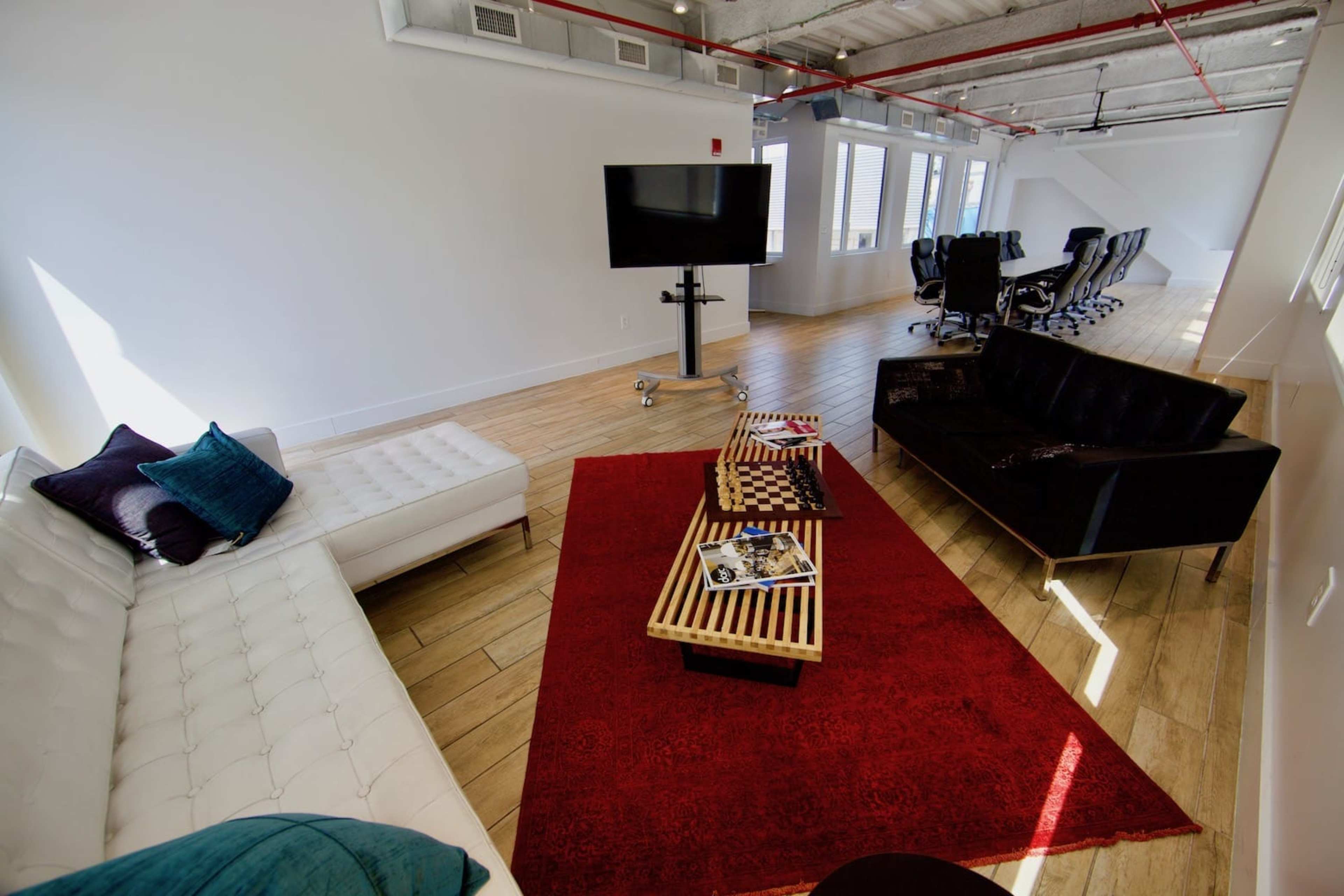 A modern office lounge area with a white sectional couch, a black couch, a red area rug, a coffee table with games, and a television.