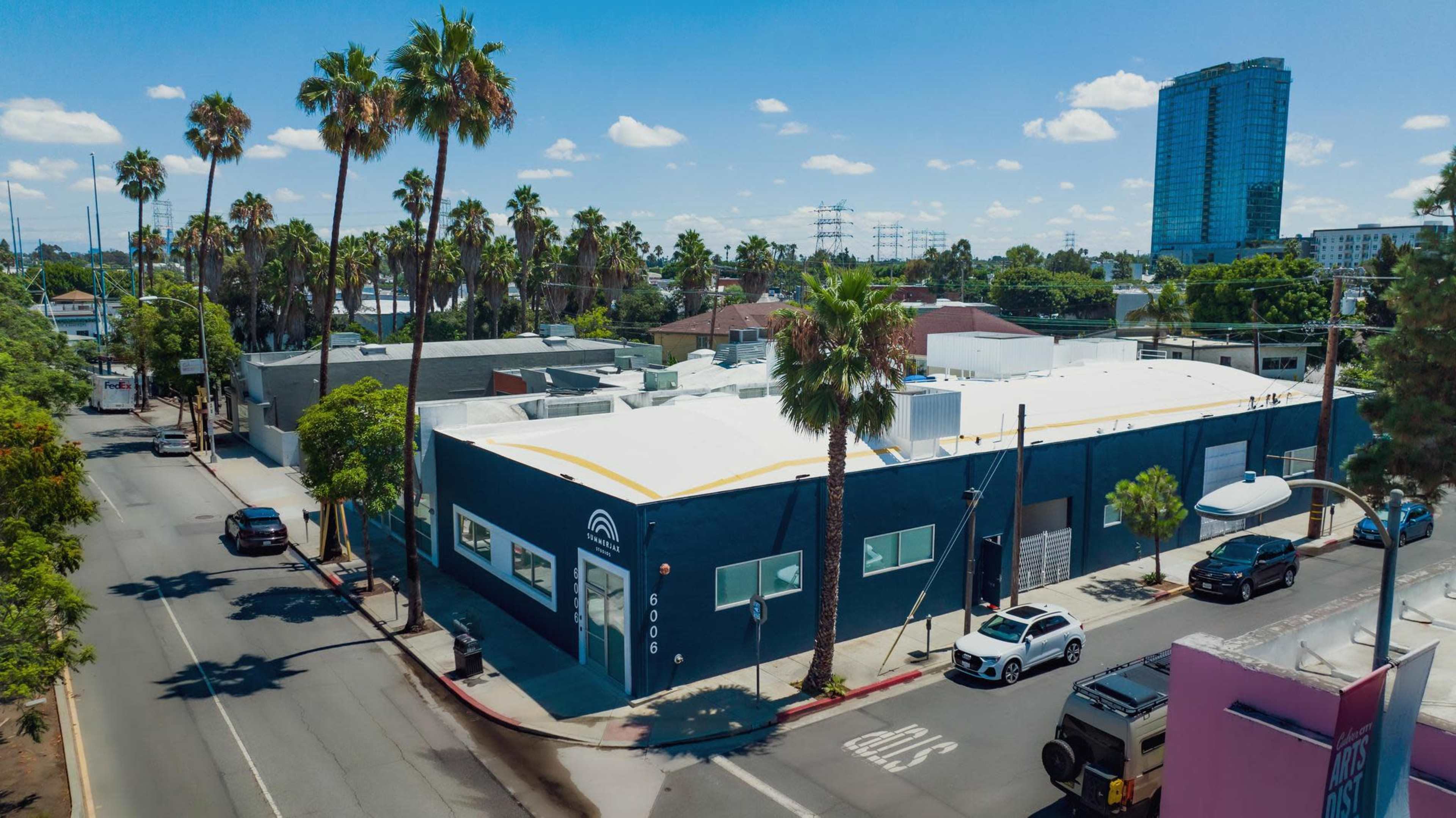 A blue commercial building with a white roof sits at the corner of a street lined with palm trees, surrounded by other low-rise structures and a tall building in the background.