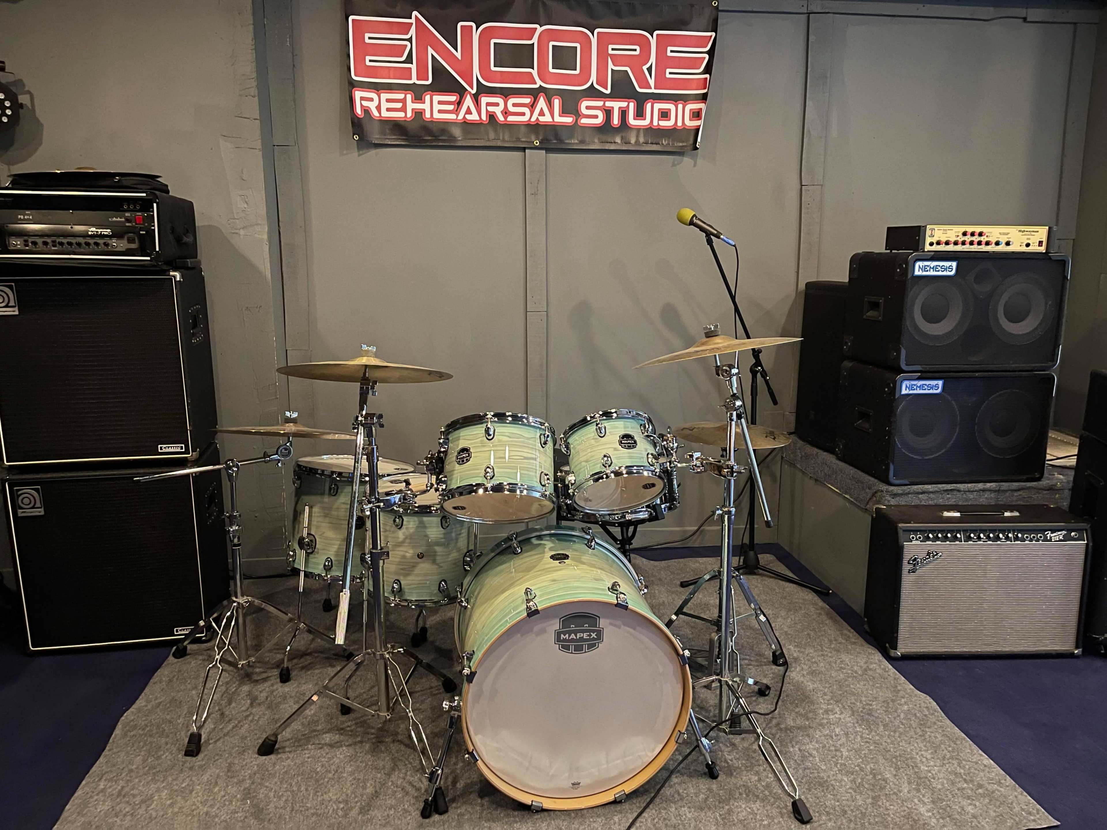 A drum set positioned in a rehearsal studio alongside amplifiers and a microphone on a stand.