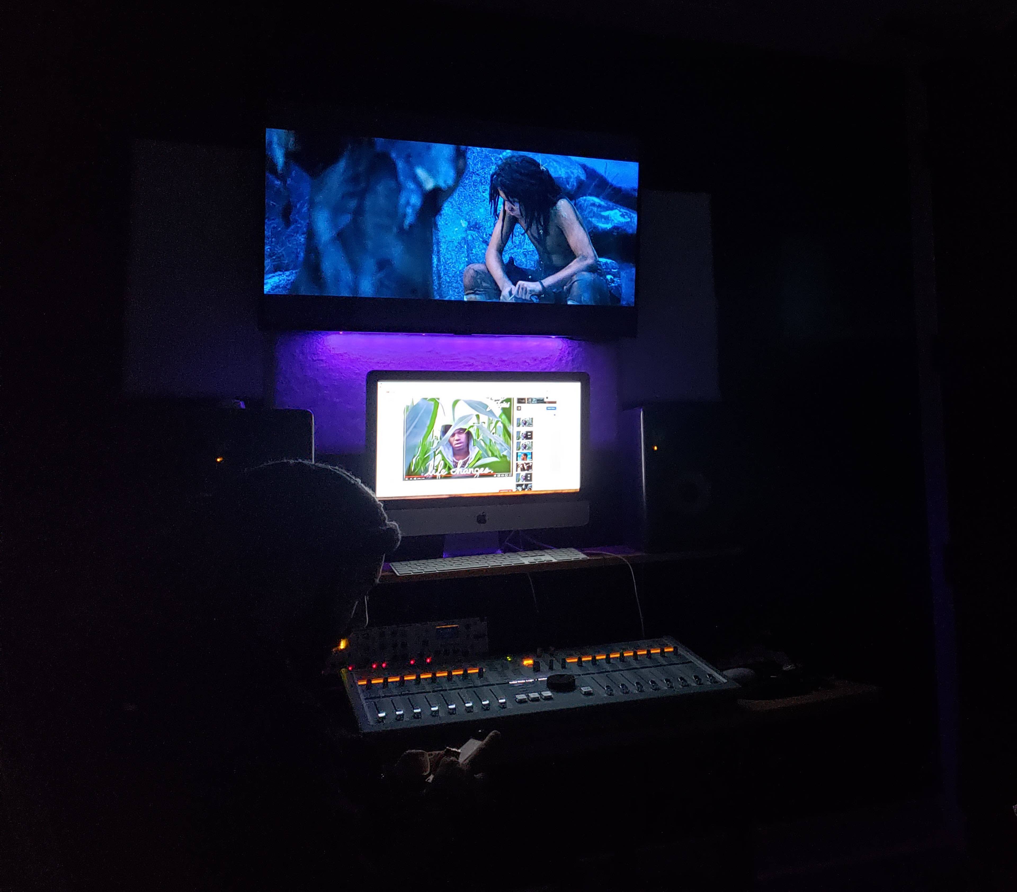 A person is seated in a dimly lit recording studio, working on a sound mixing board while a movie plays on a screen above.