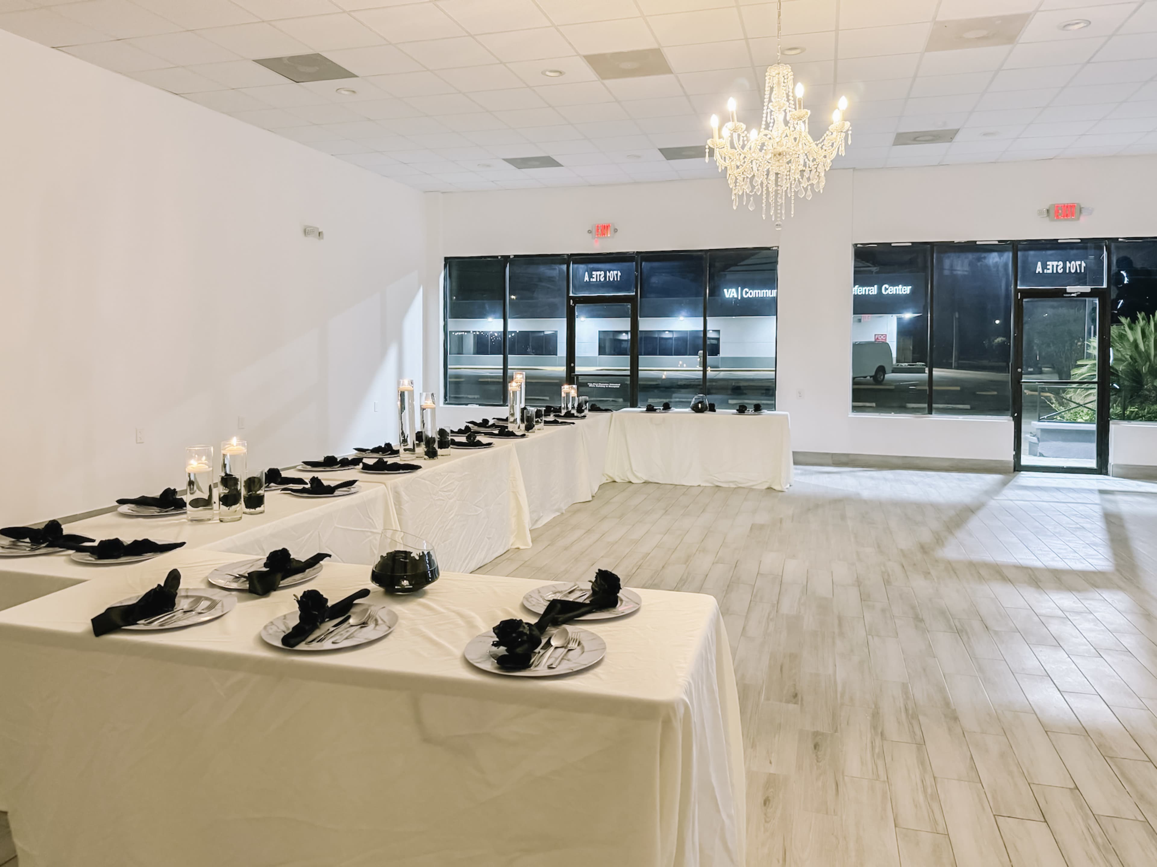 A banquet hall with two long tables covered in white cloths, set with black napkins and plates, beneath a chandelier.