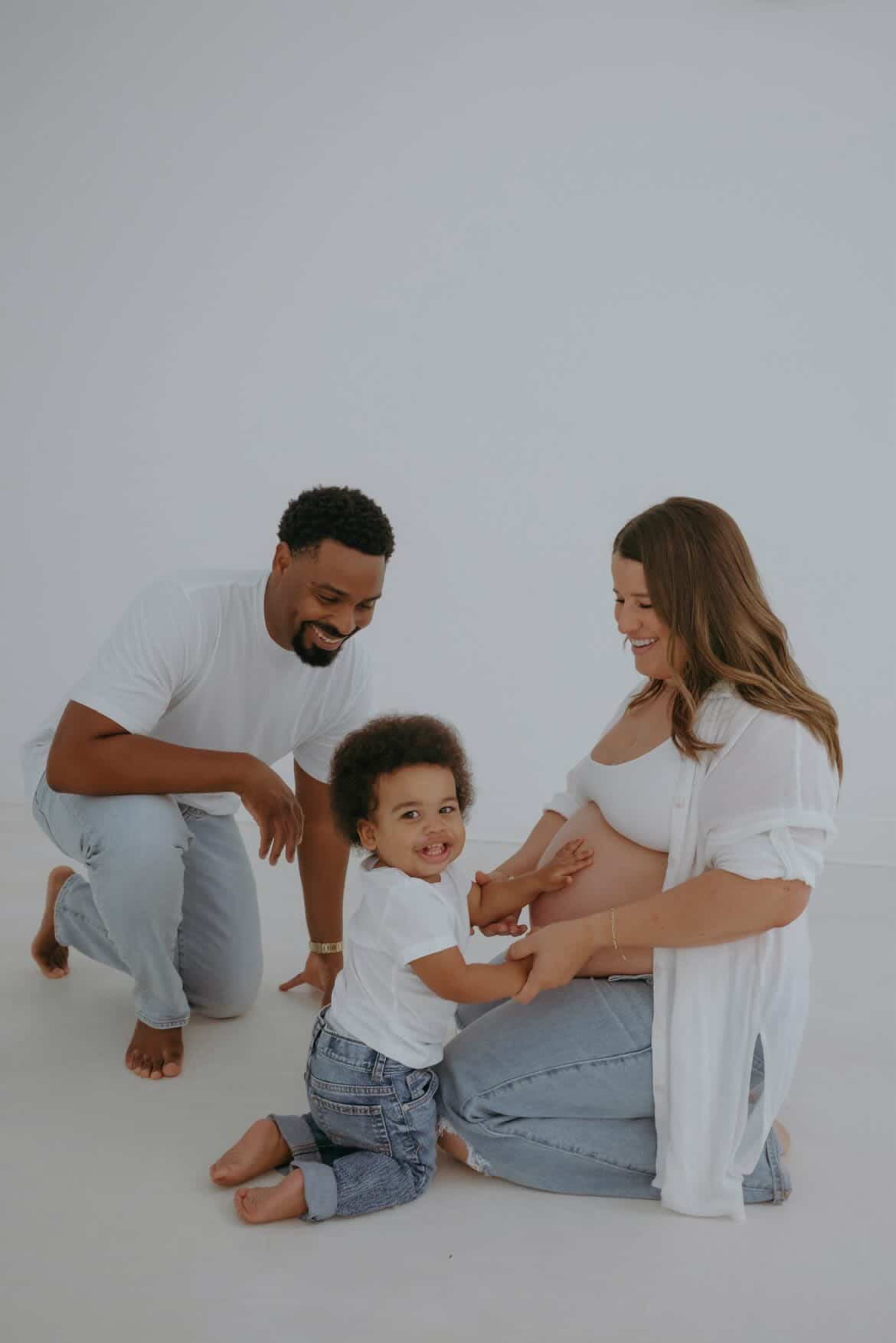 A family poses together in a studio, with a mother kneeling and smiling at her pregnant belly while a father looks on and a young child sits beside her.