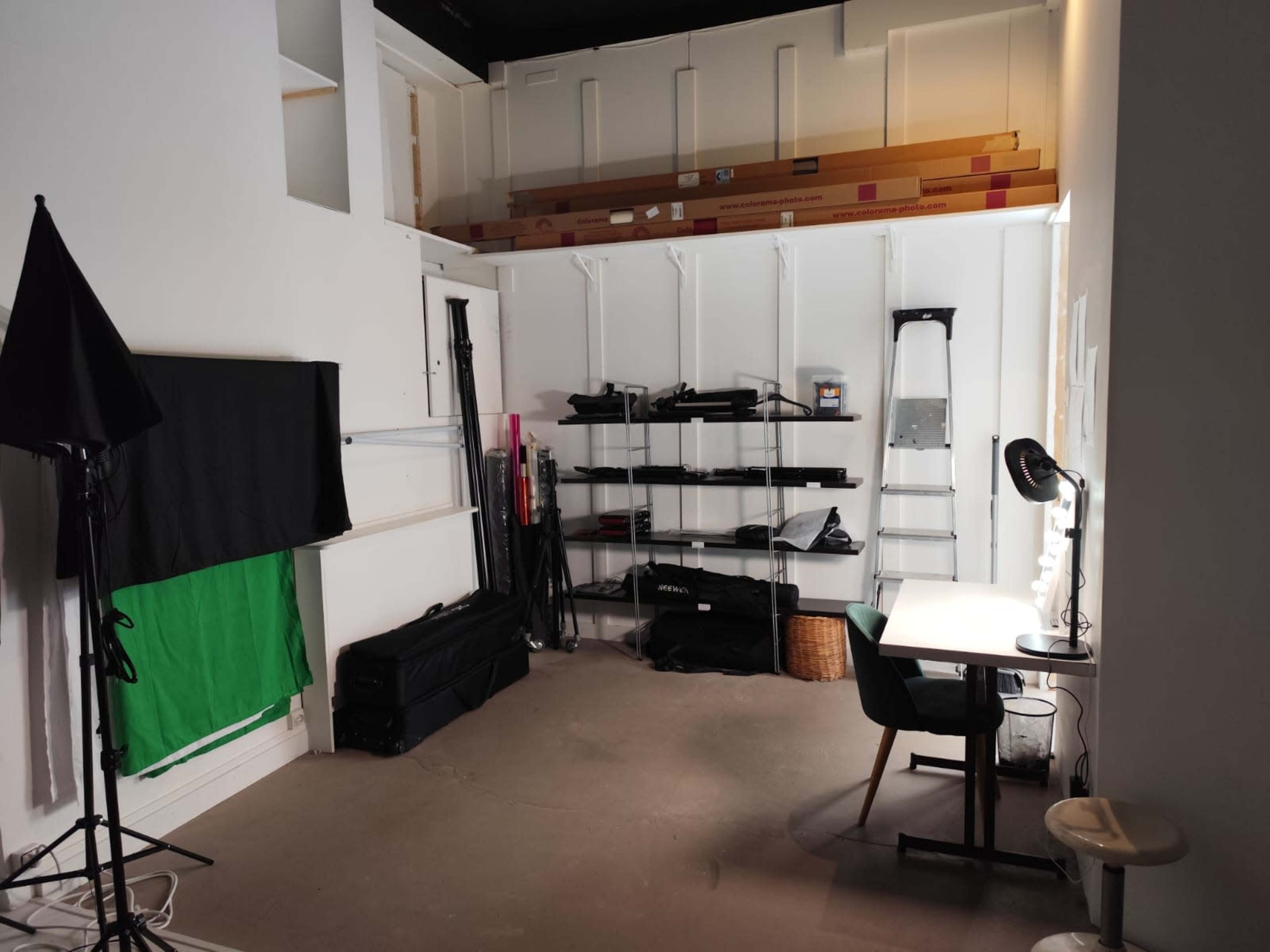 The image shows a small studio space with a green screen, shelving units filled with equipment, and a desk with a lamp and a chair.
