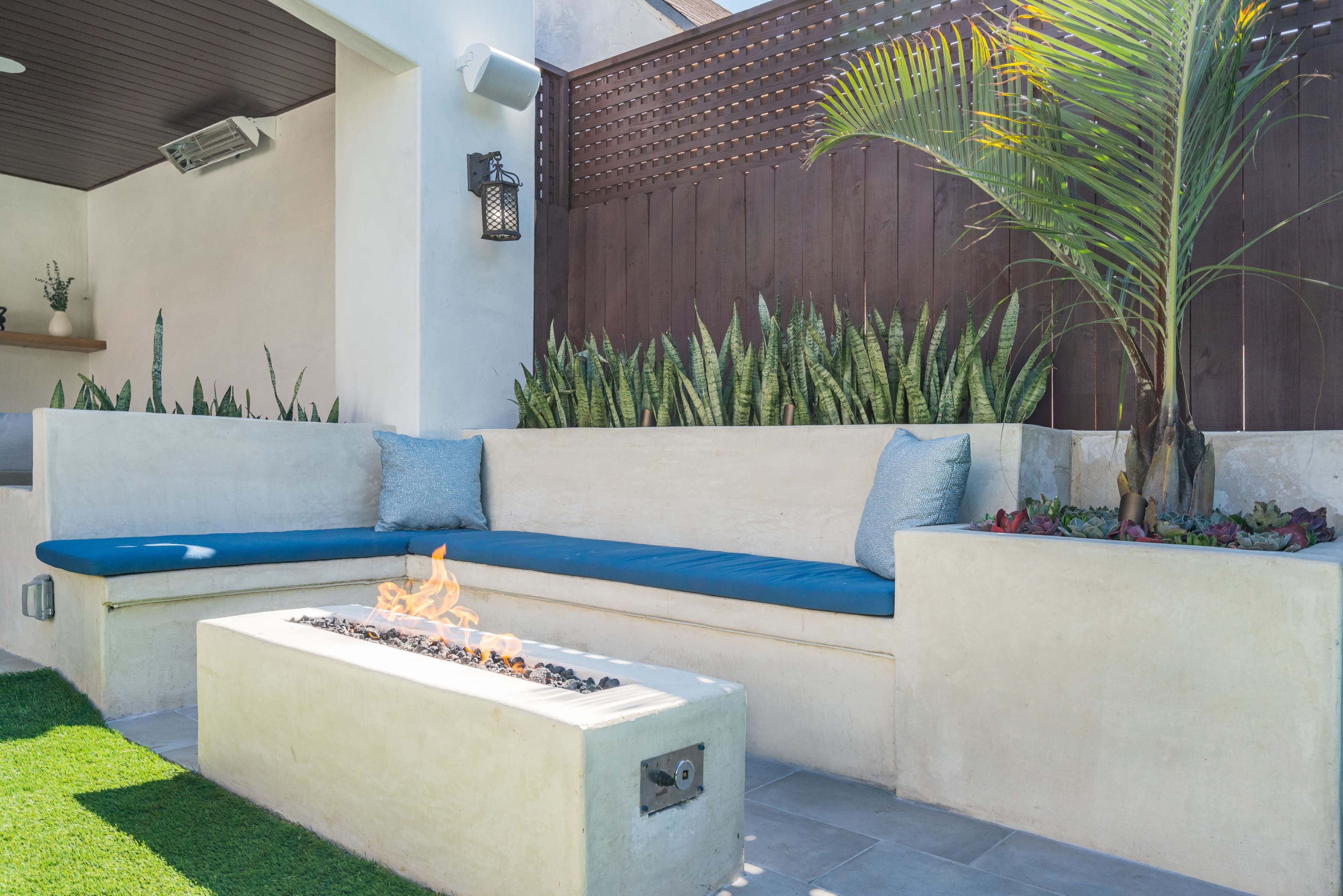 A modern outdoor seating area features a blue cushioned built-in bench adjacent to a rectangular fire pit surrounded by plants.