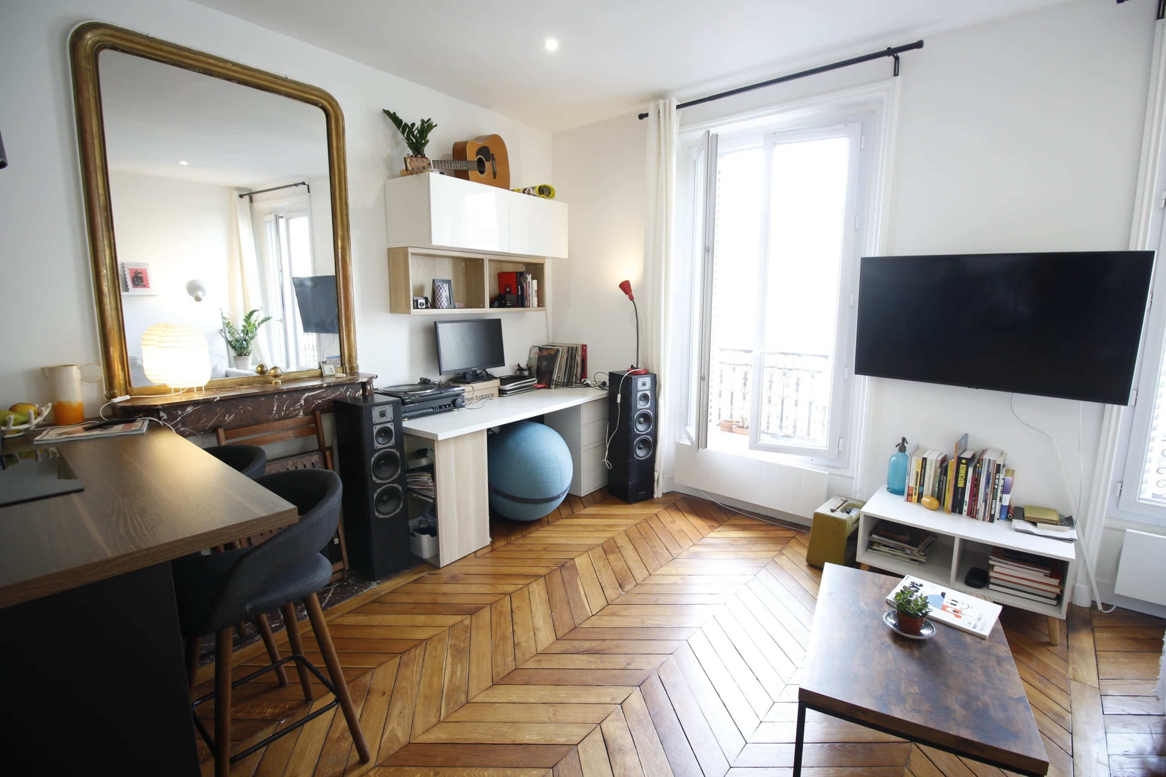 The image shows a modern living room with hardwood flooring, featuring a large mirror, a desk with computers, speakers, and a television.