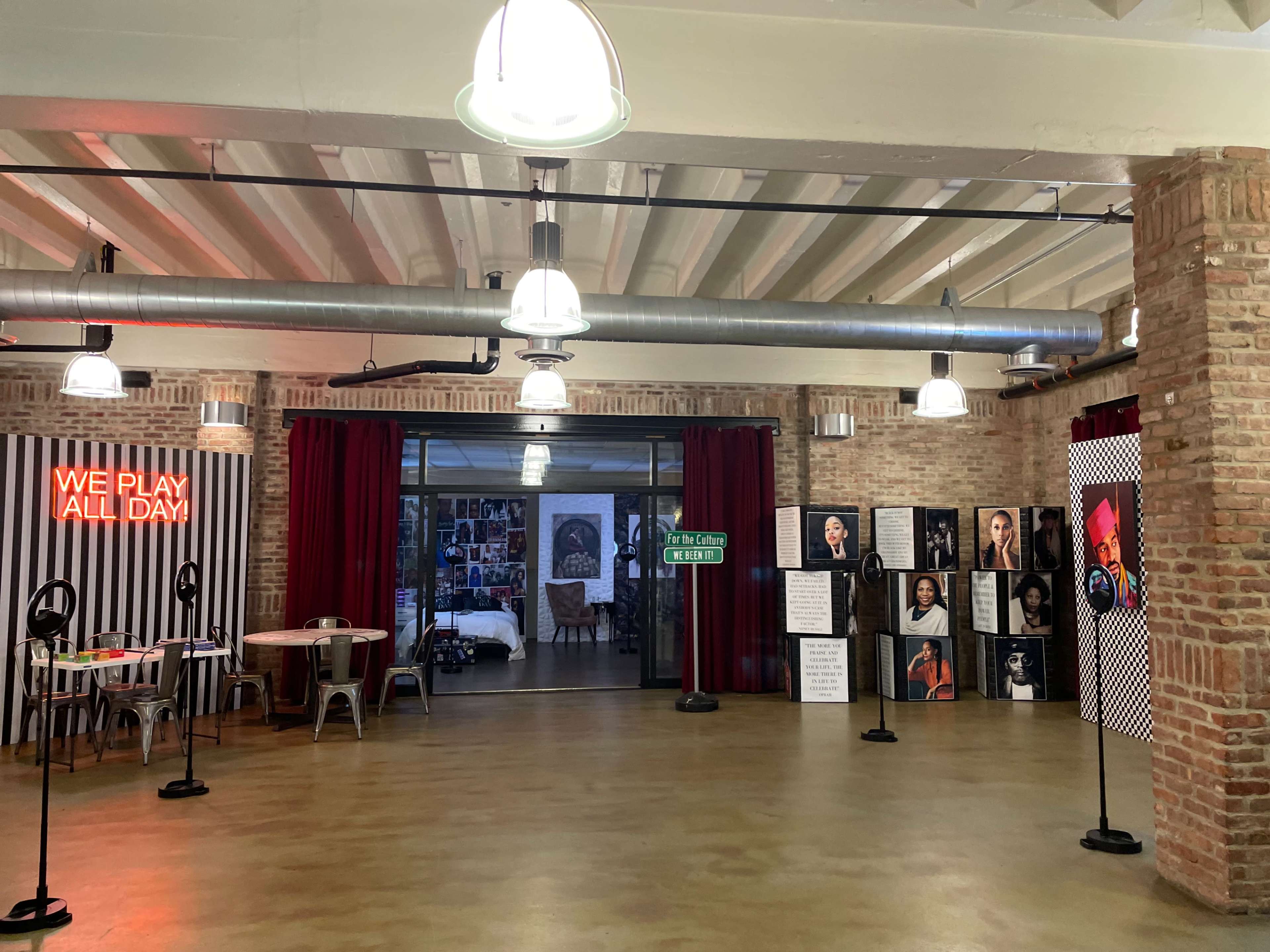 A spacious, modern room with a neon sign reading "WE PLAY ALL DAY," featuring brick walls, poster displays, and several tables and chairs.