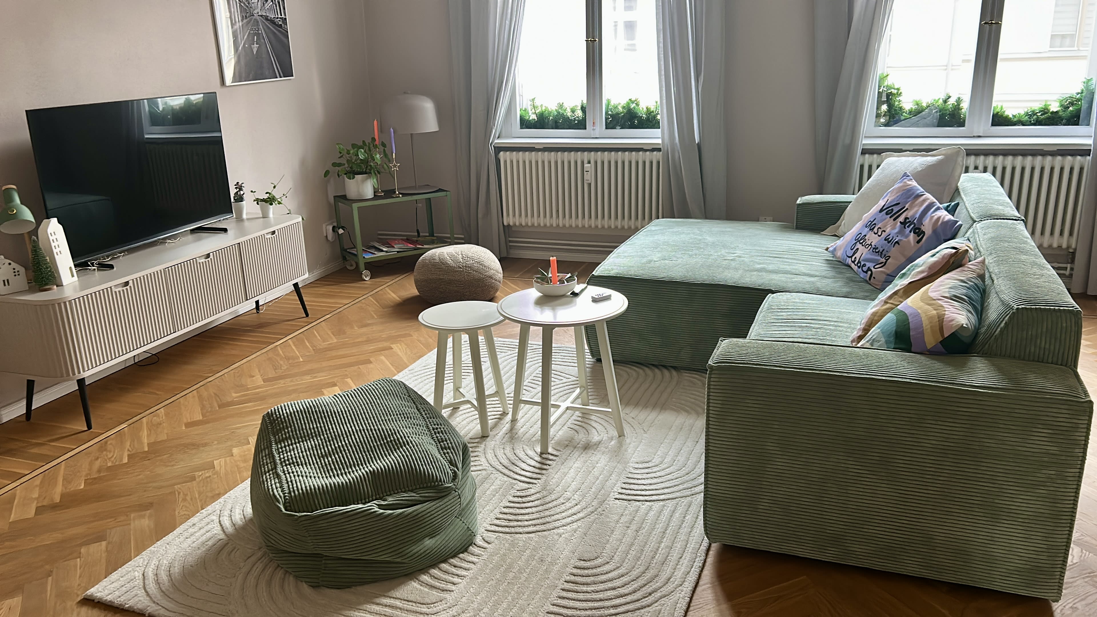 The image shows a cozy living room featuring a green sectional sofa, a television on a light-colored media console, two small tables, and a plush rug on a wooden floor.