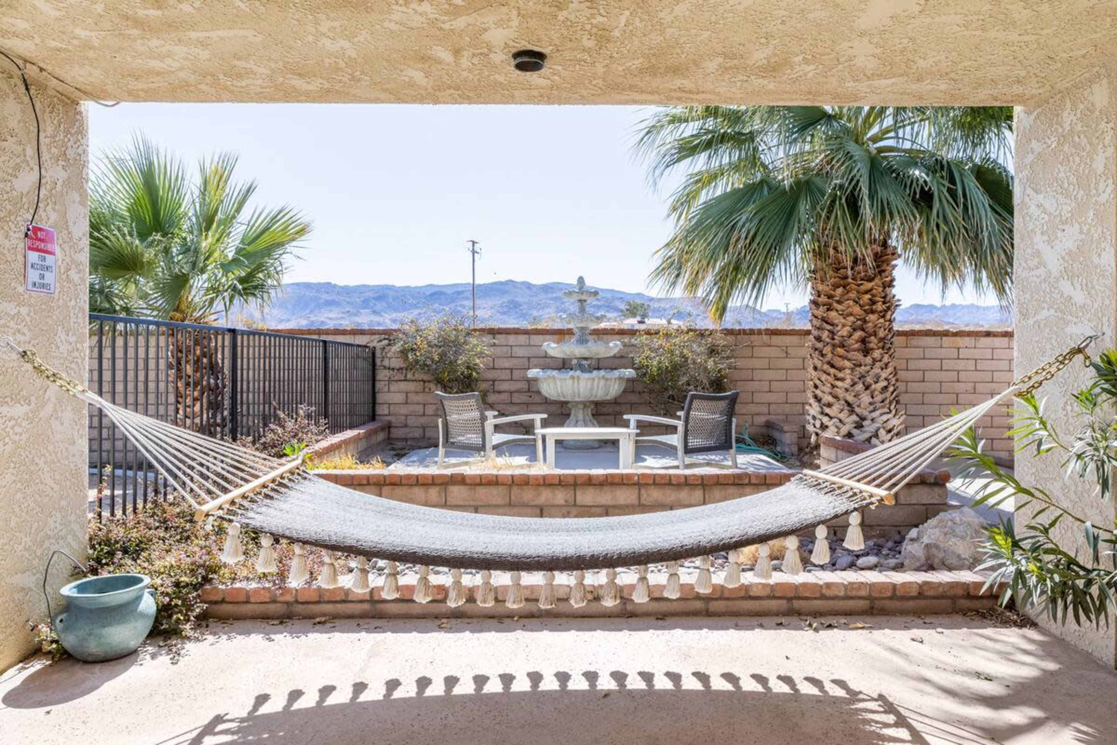 The image features a hammock set between two columns, facing a small courtyard with a fountain and seating surrounded by palm trees against a mountain backdrop.