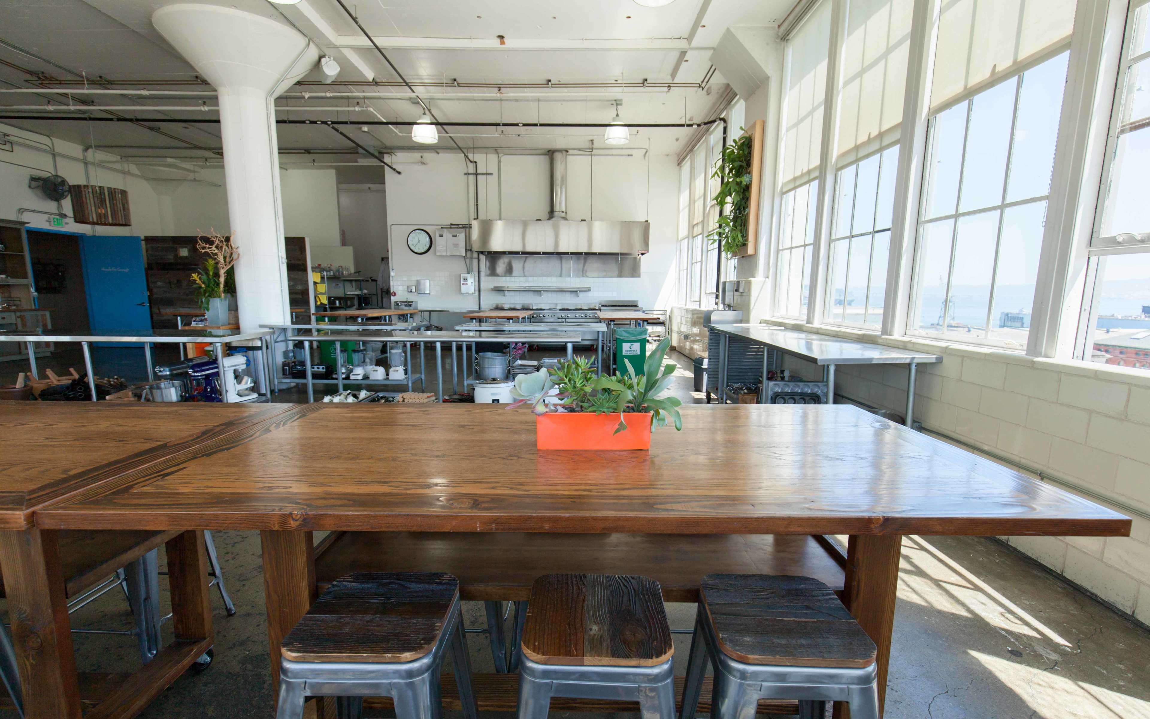 A spacious industrial kitchen with large windows, featuring a wooden dining table and metal stools, surrounded by various kitchen appliances and tools.